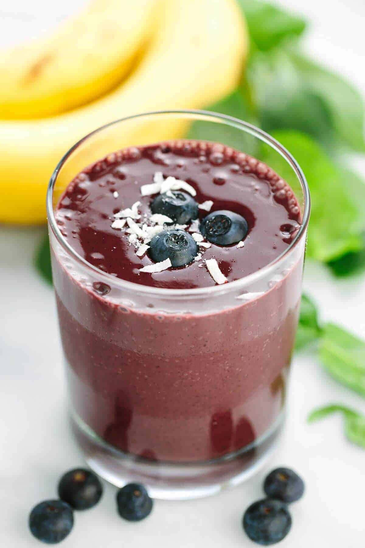 Blueberry Smoothie 桌布 壁紙 — 一杯充滿活力的紫色冰沙，上面放有藍莓和椰子片，旁邊有香蕉和薄荷葉。