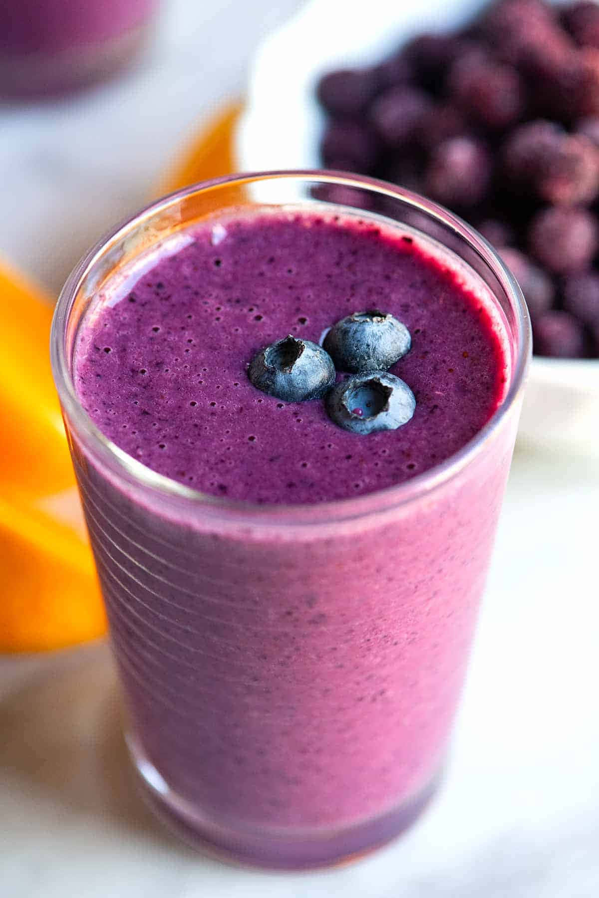 Blueberry Smoothie 桌布 壁紙 — 一杯充滿活力的紫色冰沙，上面放著新鮮藍莓，旁邊有一堆藍莓和橙片。
