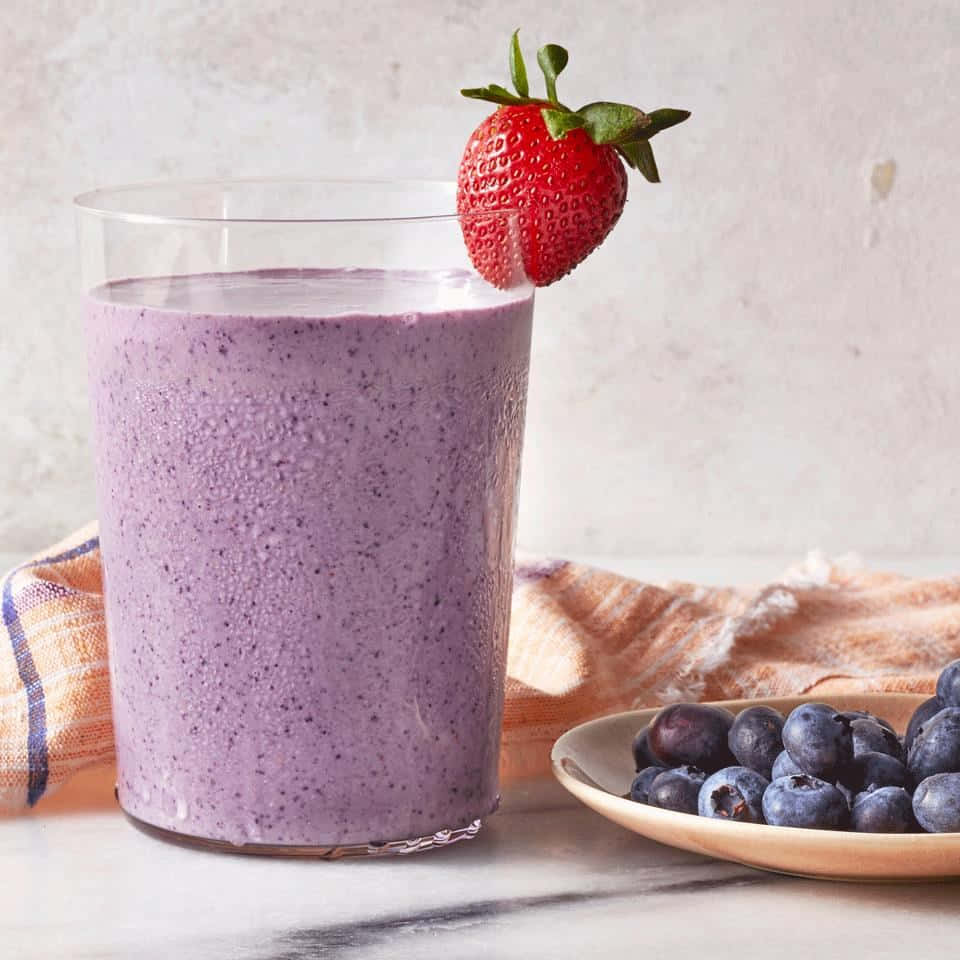 Blueberry Smoothie 桌布 壁紙 — 一杯充滿活力的紫色冰沙，用新鮮草莓裝飾，旁邊放著一碗藍莓，置於大理石表面上。
