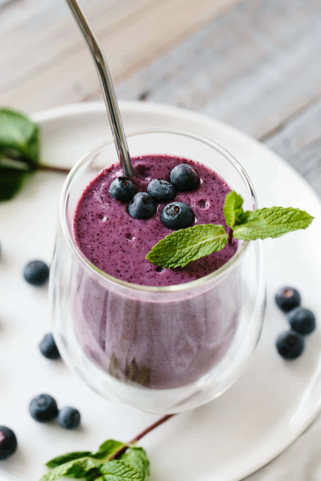 Blueberry Smoothie 桌布 壁紙 — 一杯充滿活力的紫色藍莓冰沙，上面放上新鮮藍莓和薄荷葉，置於白色碟子上。