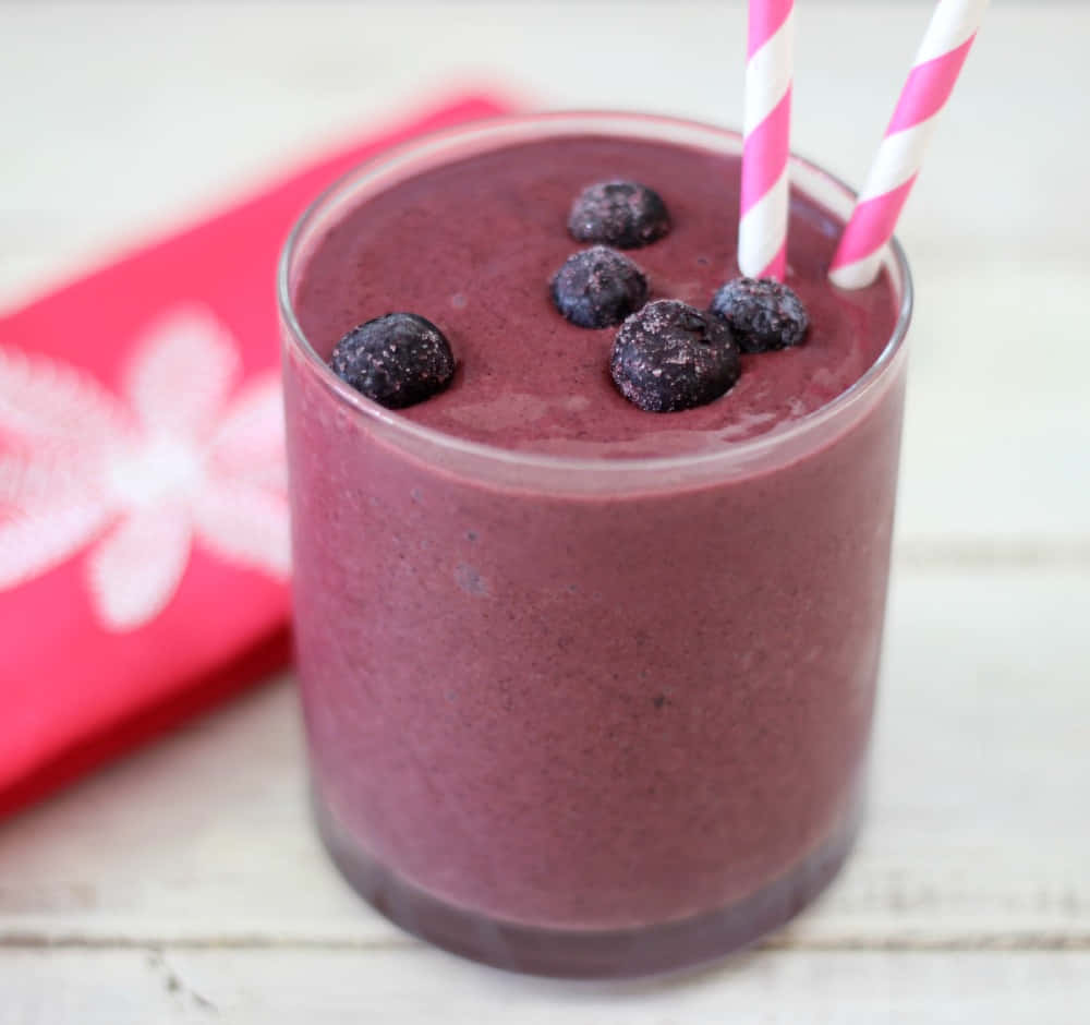 Blueberry Smoothie 桌布 壁紙 — 一杯充滿活力的紫色藍莓奶昔，上面放上新鮮莓果和粉紅條紋飲管，放在白色木質表面上，旁邊有一張紅色花卉圖案餐巾。