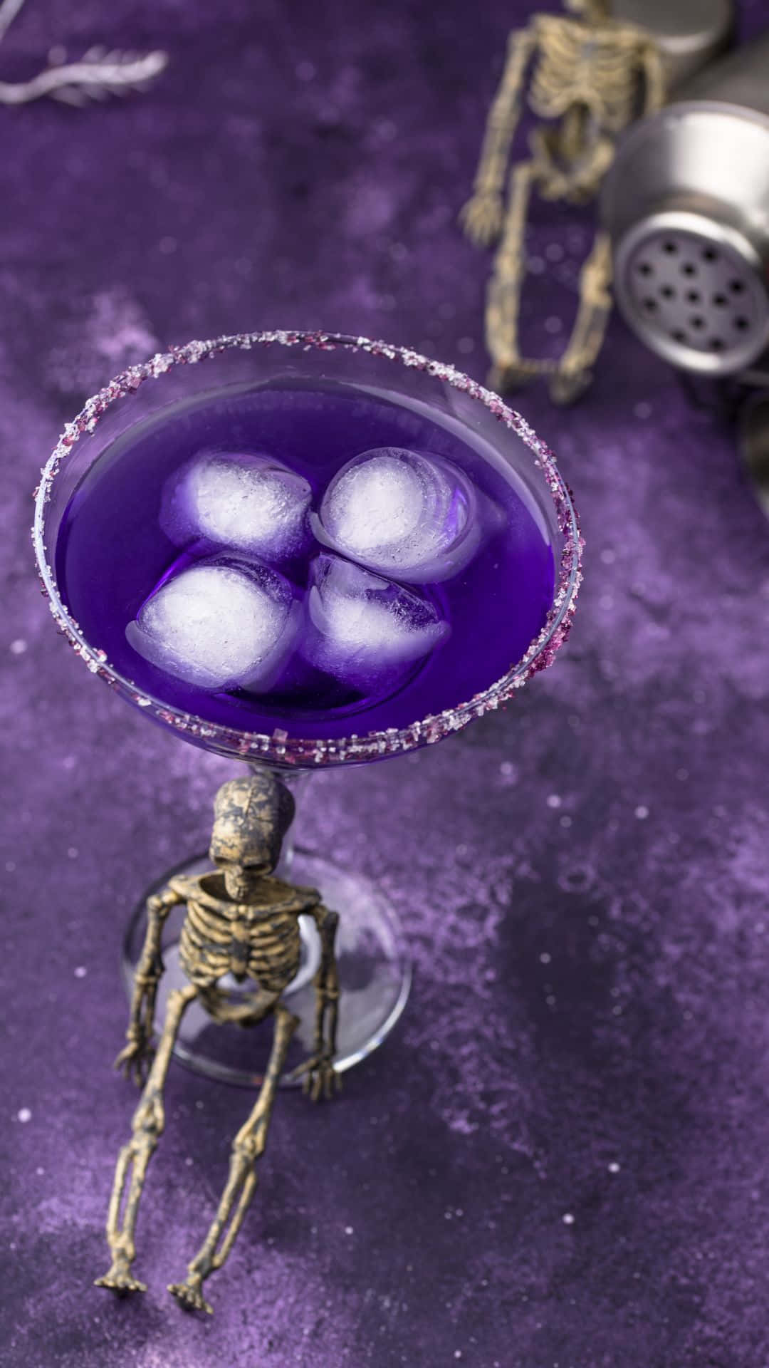 Spooky Cocktails 桌布 壁紙 — 一杯充滿活力的紫色雞尾酒，帶有閃粉邊緣和冰塊，放在一個由小型骷髏公仔手持的馬提尼杯中，置於相配的紫色紋理表面上。