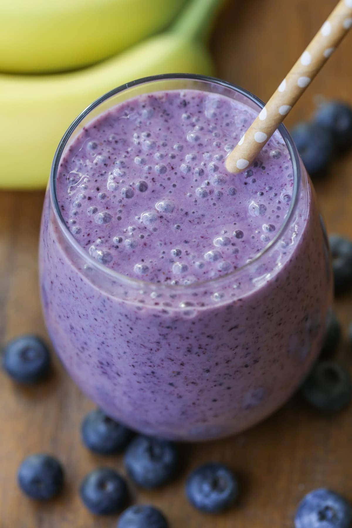 Blueberry Smoothie 桌布 壁紙 — 一杯充滿活力的紫藍色藍莓香蕉冰沙，放在玻璃杯中，插著波點飲管，周圍是新鮮藍莓和香蕉，置於木質表面上。