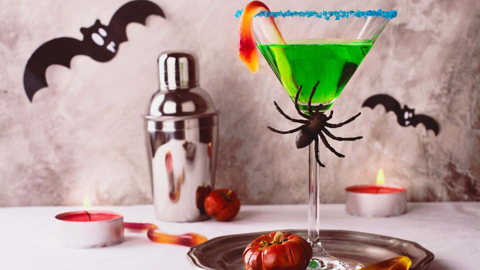 Spooky Cocktails 桌布 壁紙 — 一杯充滿活力的綠色萬聖節雞尾酒，盛載於馬提尼杯中，飾有藍色邊糖果蜘蛛和橙色糖果裝飾。