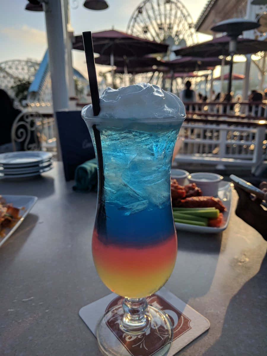 Beautiful Drink 桌布 壁紙 — 一杯充滿活力的藍、紅、黃分層雞尾酒，配上忌廉和吸管，放在主題公園戶外庭院的杯墊上，背景有摩天輪。