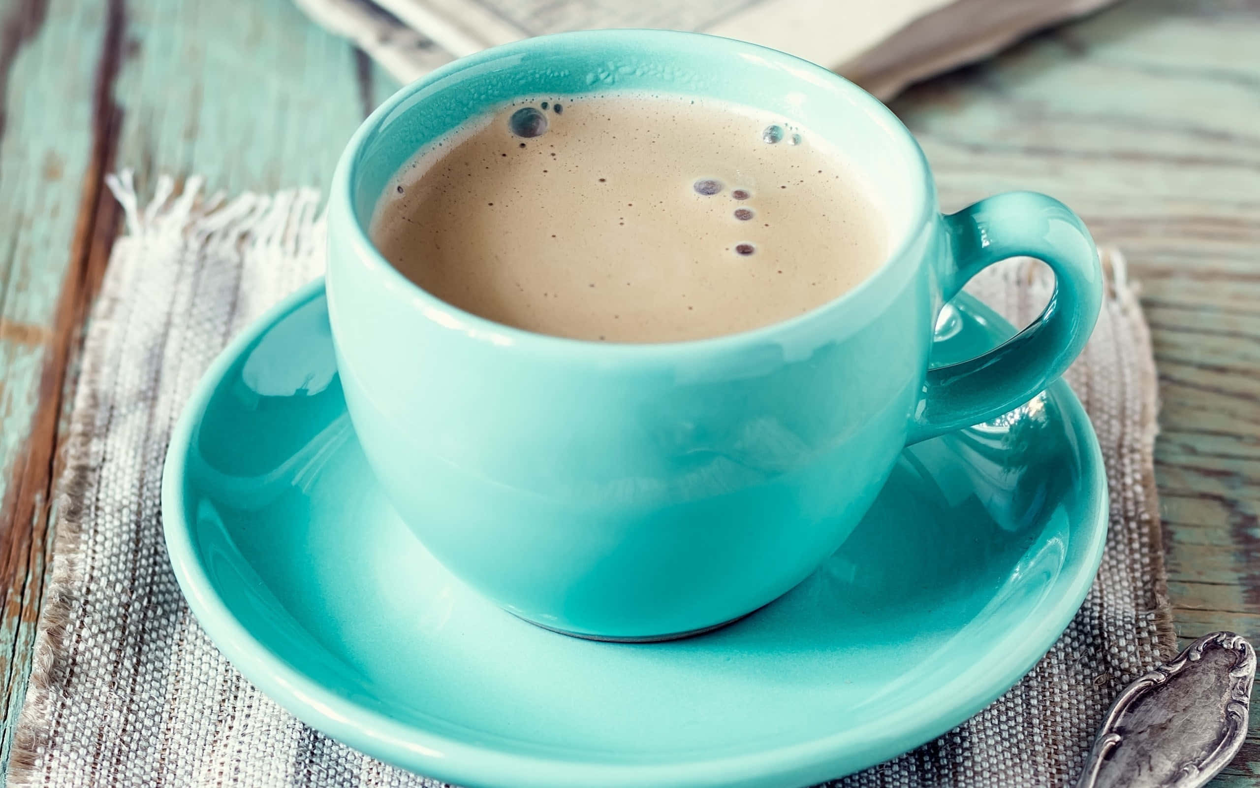 Coffee Cup 桌布 壁紙 — 一杯充滿活力的藍綠色泡沫咖啡放在相配的碟子上，置於一張質樸的木桌上，旁邊有一塊有紋理的布和一把復古湯匙。