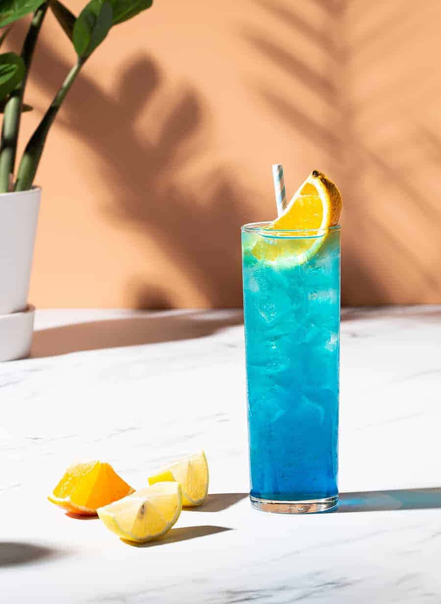 Beautiful Drink 桌布 壁紙 — 一杯充滿活力的藍色雞尾酒，加冰和橙片，放在大理石桌上，旁邊有柑橘楔形塊，沐浴在溫暖的陽光下。