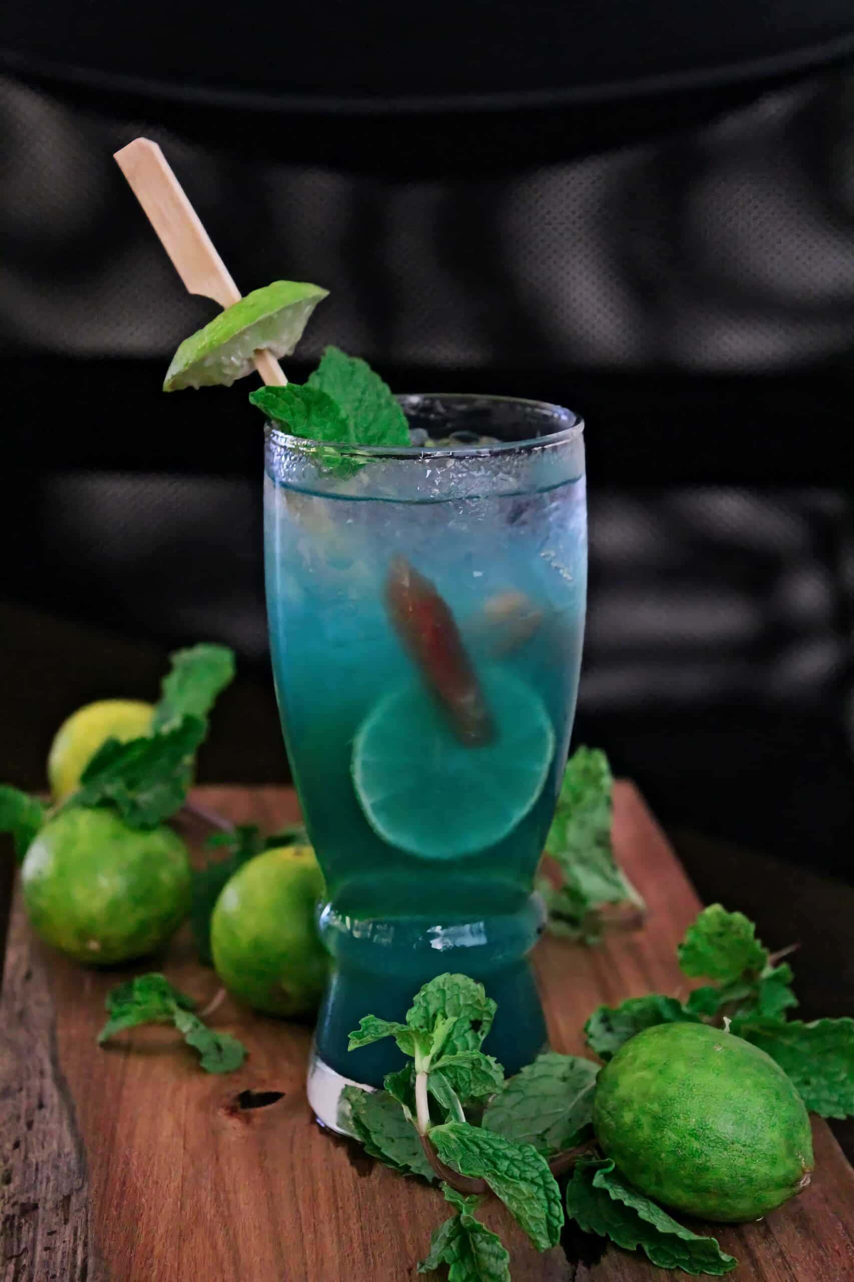 Beautiful Drink 桌布 壁紙 — 一杯充滿活力的藍色雞尾酒，飾以青檸和薄荷，放在木板上，周圍是新鮮的青檸和薄荷葉。