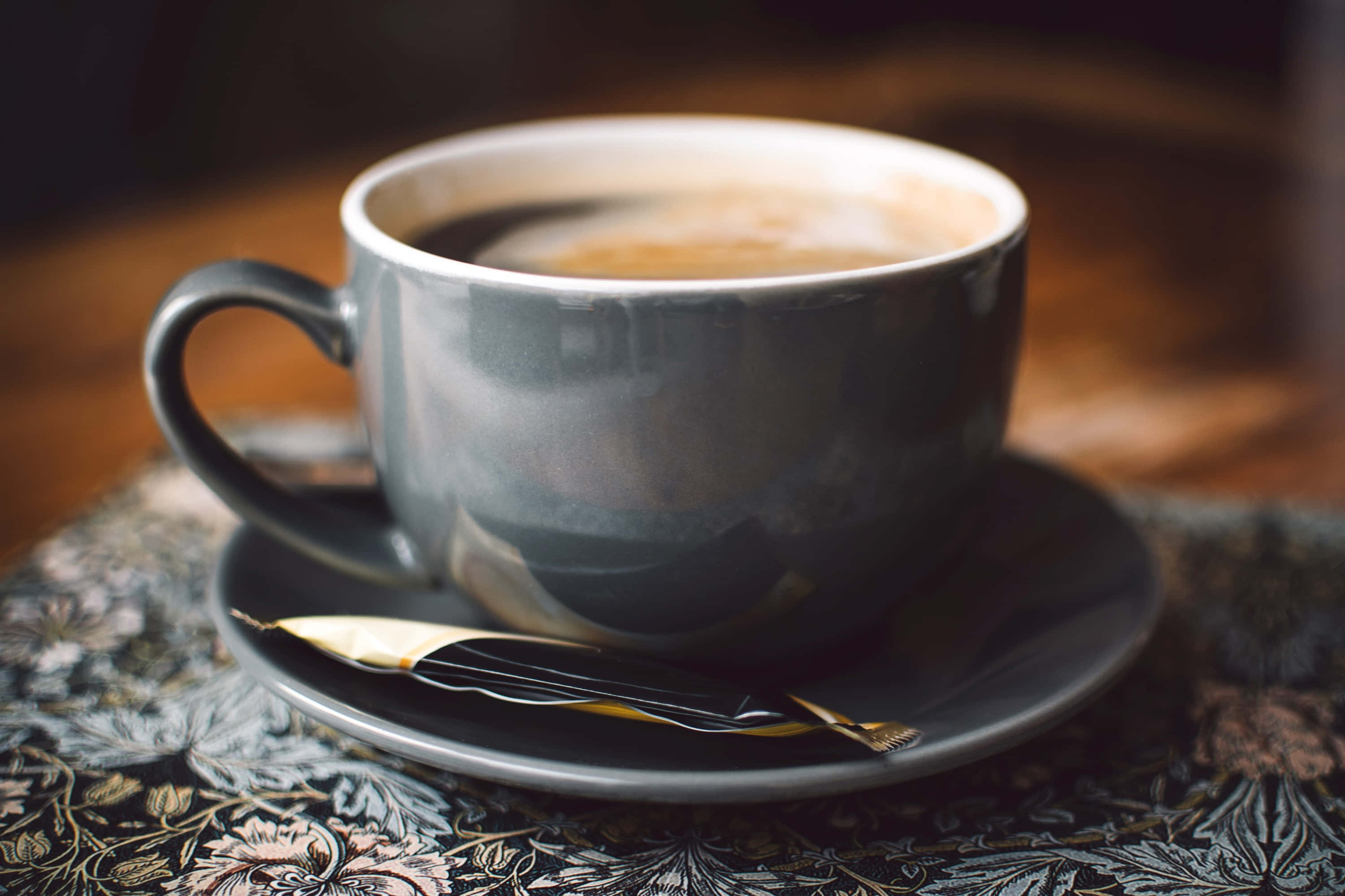 Coffee Cup 桌布 壁紙 — 一杯冒煙的咖啡放在灰色陶瓷杯裡，擱在碟子上，旁邊有一包糖，底下是花卉圖案的桌布。