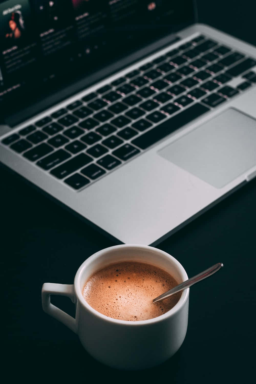 Minimalist Coffee 桌布 壁紙 — 一杯冒煙的咖啡配上一隻匙羹，放在昏暗的桌子上，旁邊是一部手提電腦，在微光中柔和地發光。