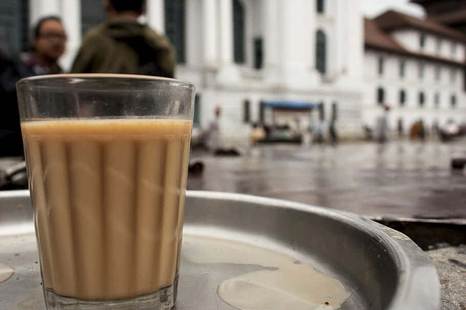 Milk And Coffee 桌布 壁紙 — 一杯冒煙的奶茶放在金屬托盤上，背景是模糊的濕漉街道和殖民風格建築。