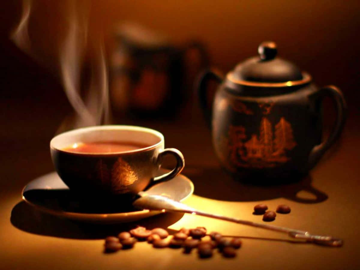 Coffee Cup 桌布 壁紙 — 一杯冒煙的茶放在一個華麗的茶壺旁邊，散落的咖啡豆在溫暖的金色燈光下。