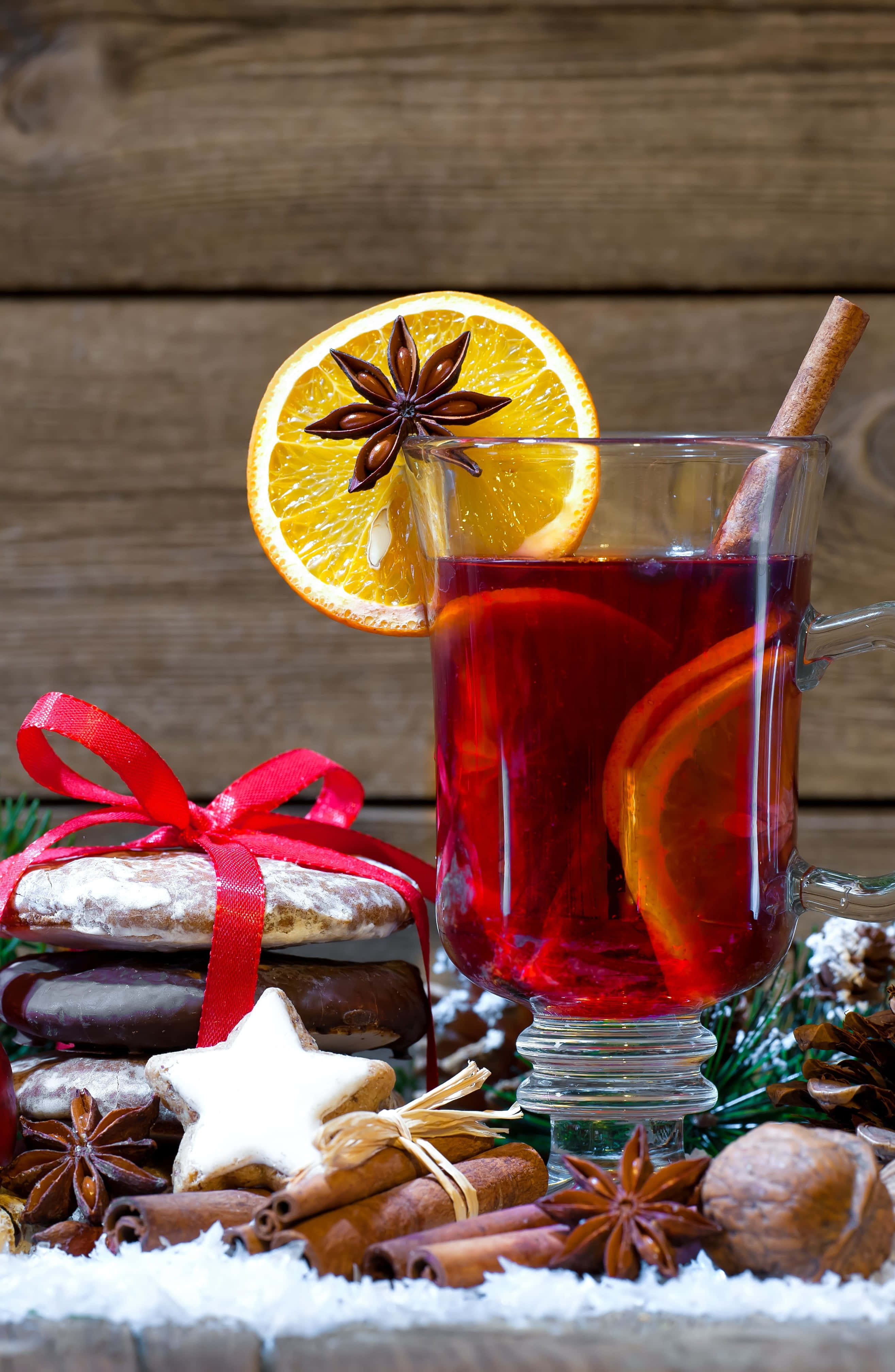 Mulled Wine 桌布 壁紙 — 一杯冒煙的香料熱紅酒，飾以橙片、八角及肉桂棒，放在雪覆蓋的木頭上，旁邊有節日餅乾和松果。