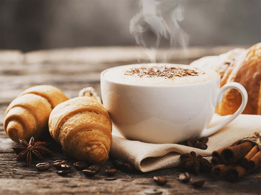 Milk And Coffee 桌布 壁紙 — 一杯冒著熱氣的咖啡，配上肉桂和八角，放在鄉村風木桌上，旁邊是金黃色的牛角包。