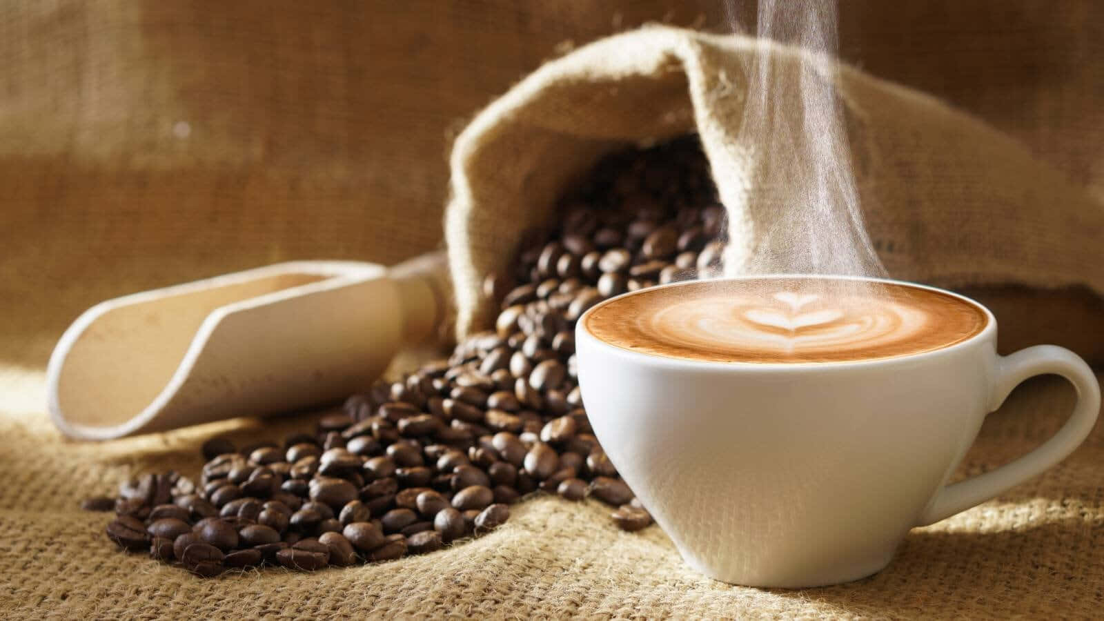 Brown Coffee 桌布 壁紙 — 一杯冒著熱氣的拉花咖啡旁邊，散落著從麻布袋中灑出的咖啡豆，置於有紋理的布料上。