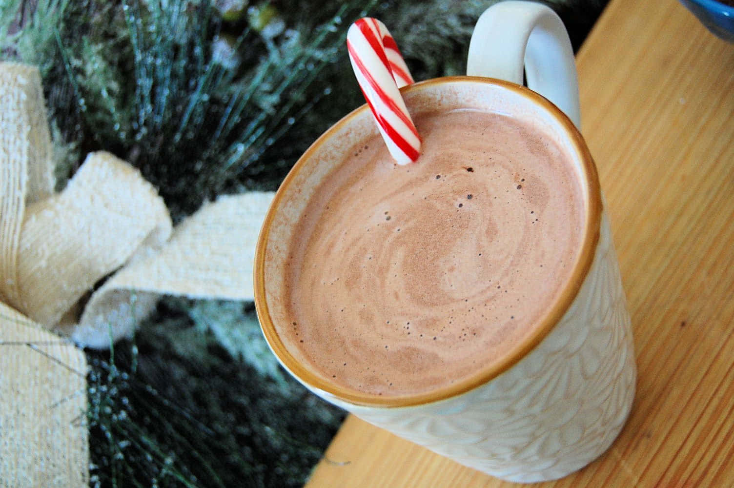 Hot Chocolate 桌布 壁紙 — 一杯冒著熱氣的濃郁朱古力熱可可，配上一根拐杖糖攪拌棒，放在木桌上，旁邊有節日綠植和一條奶油色絲帶。