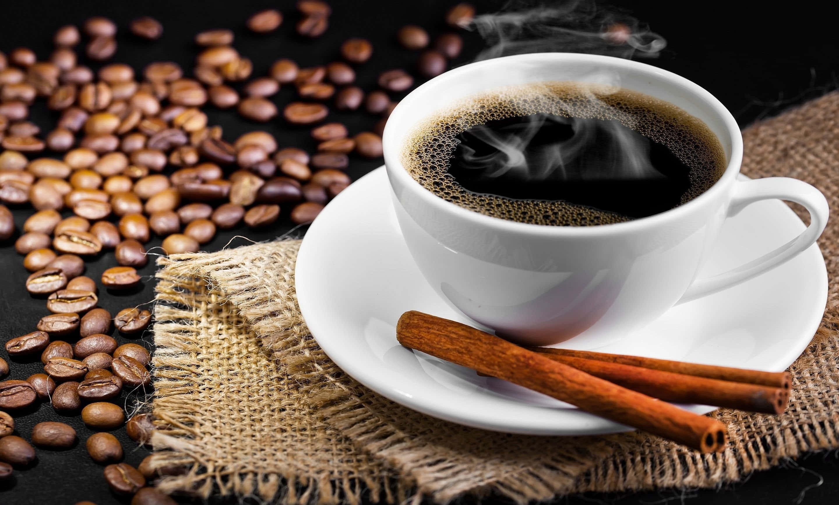 Black Coffee 桌布 壁紙 — 一杯冒著熱氣的黑咖啡配以肉桂棒，放在麻布上的碟子裡，周圍散落著烘焙咖啡豆在深色表面上。