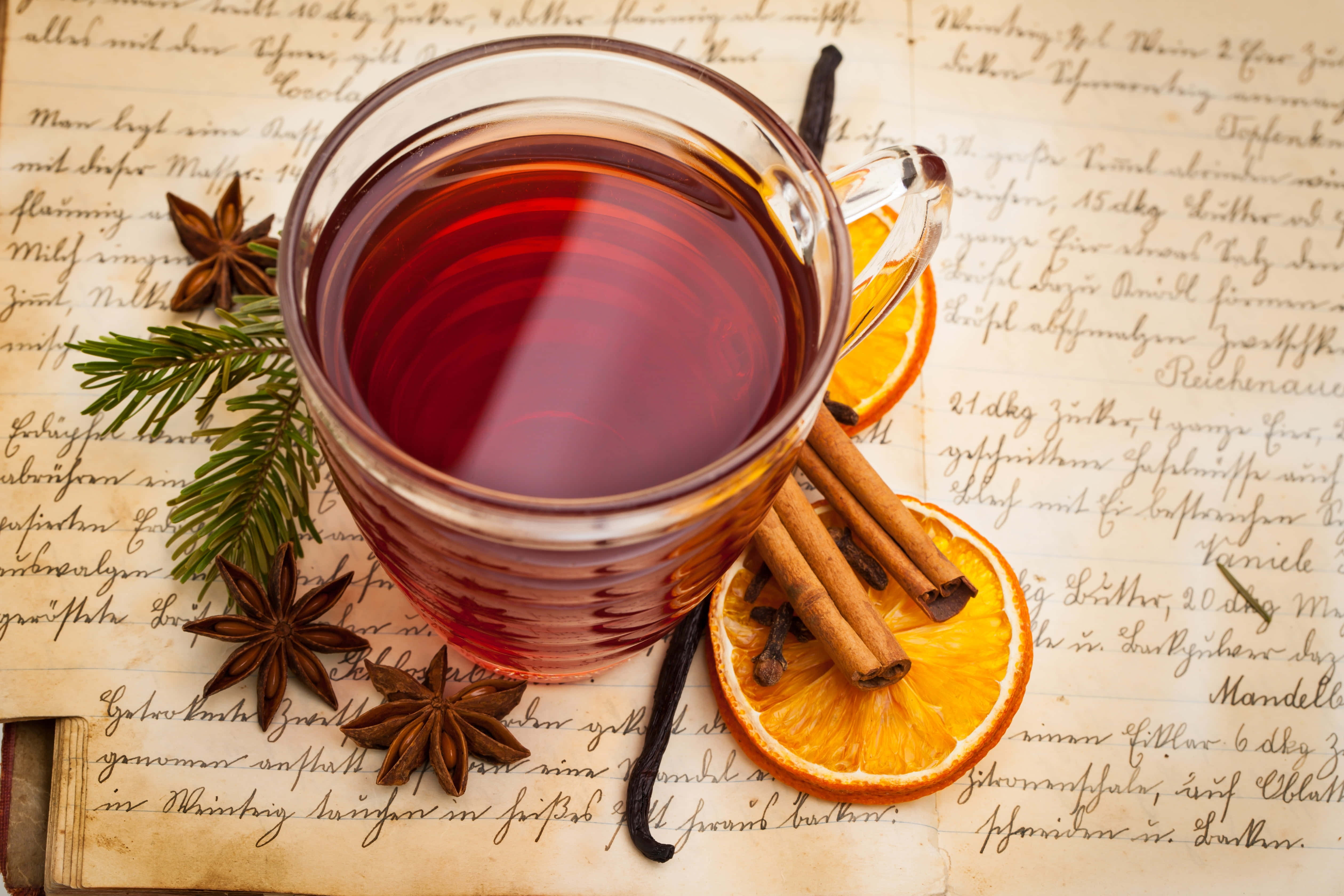 Mulled Wine 桌布 壁紙 — 一杯冒著蒸氣的香料茶玻璃杯放在復古手寫紙上，周圍有肉桂棒、八角、橙片和雲呢拿豆。