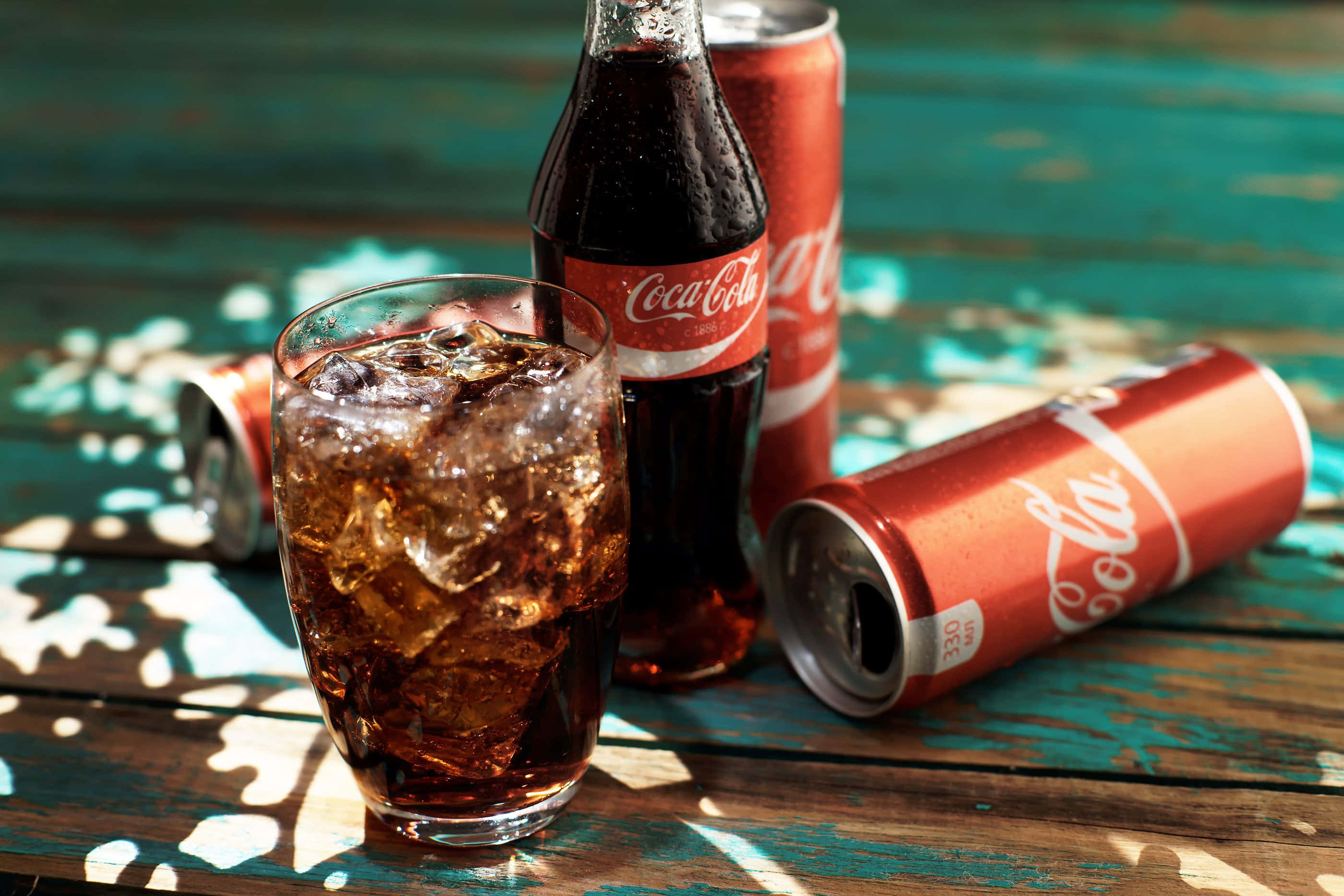 coca cola 桌布 壁紙 — 一杯冰凍可口可樂放在一個風化的藍綠色木桌上，旁邊有一個瓶子和幾個罐子。