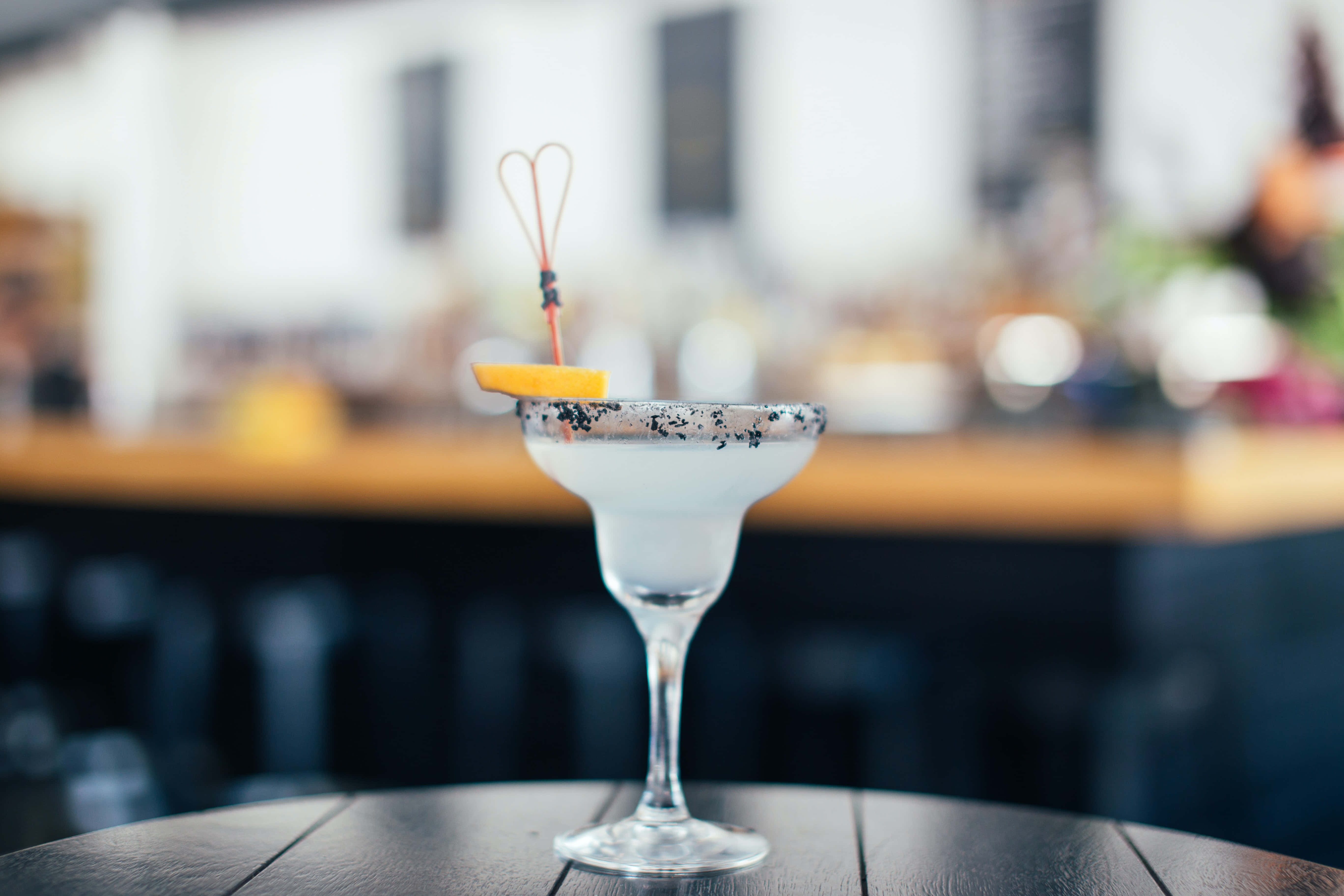 Summer Drinks 桌布 壁紙 — 一杯冰凍的瑪格麗特雞尾酒，帶有鹽邊和橙片裝飾，放在深色木桌上，背景是柔和模糊的酒吧環境。