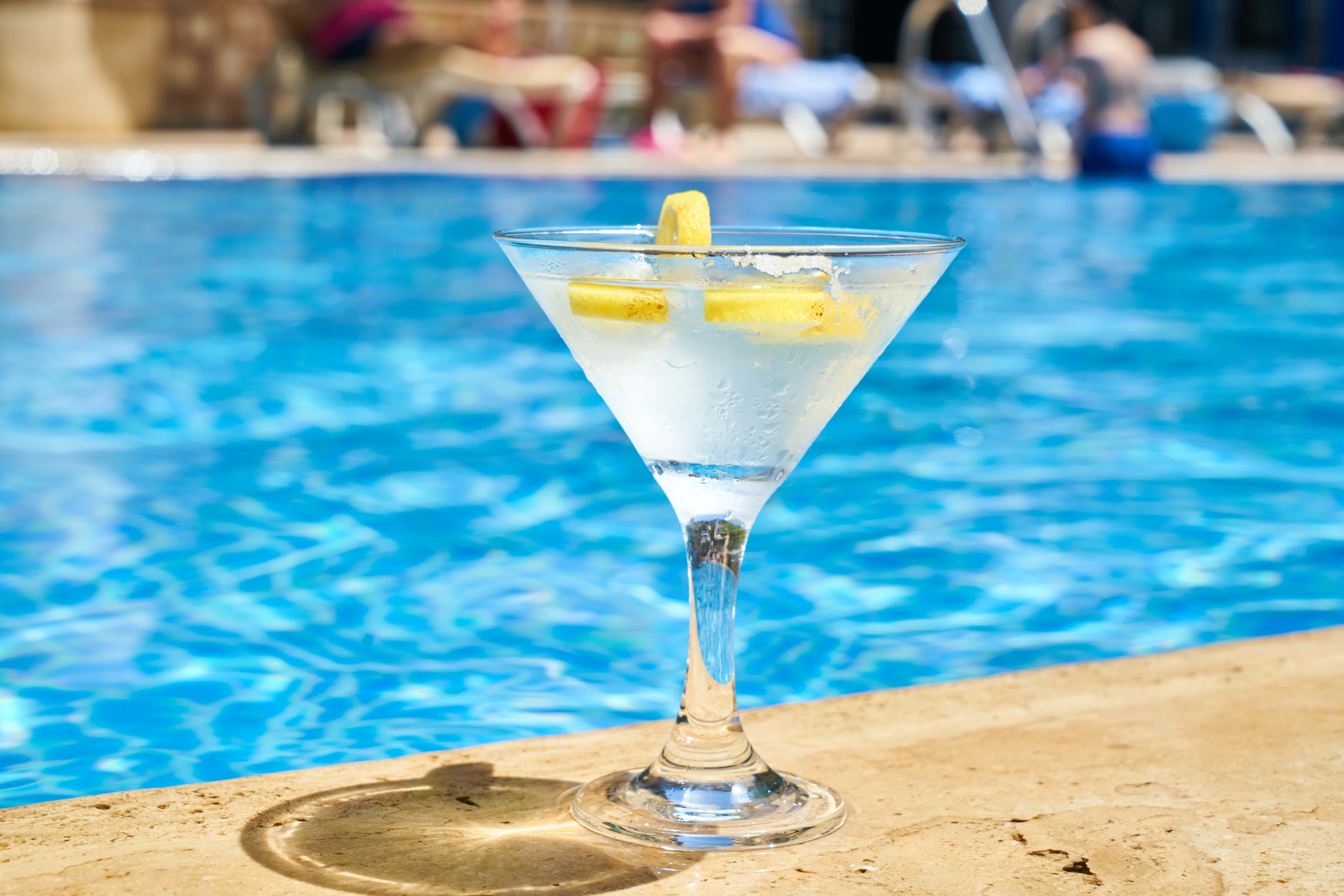 Summer Drinks 桌布 壁紙 — 一杯冰凍馬天尼配檸檬裝飾，放在陽光普照下閃爍藍色水邊的泳池石上。
