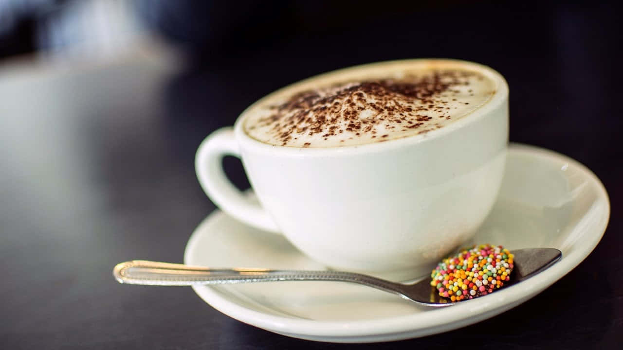 Milk And Coffee 桌布 壁紙 — 一杯奶油卡布奇諾，上面撒上可可粉和彩色糖粒，放在碟子上，旁邊有一隻匙羹。