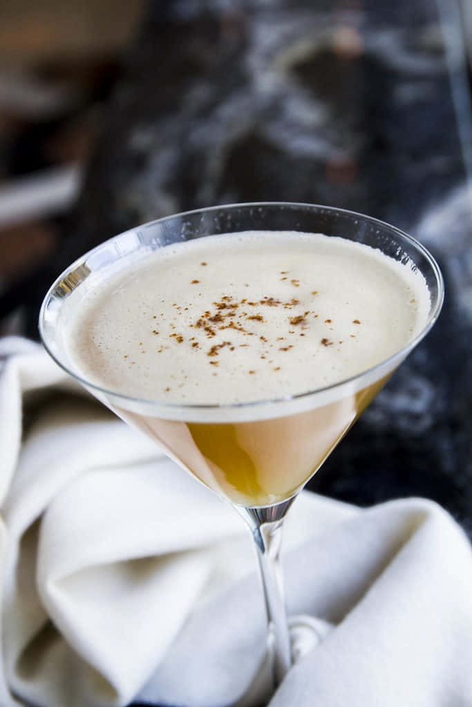 Winter Drinks 桌布 壁紙 — 一杯奶油雞尾酒盛載於馬提尼杯中，以香料點綴，優雅地擺放在白色布料上，背景為深色大理石。