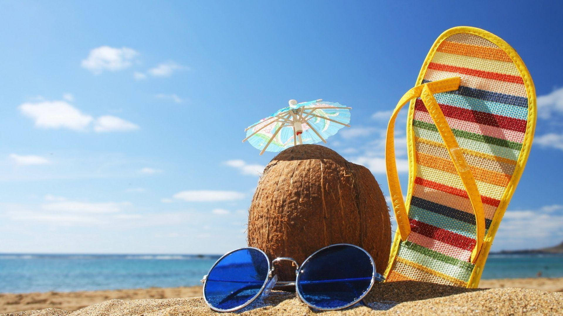 Summer Beach 桌布 壁紙 — 一杯帶有太陽傘、太陽眼鏡和條紋人字拖的椰子飲品，放在陽光普照的沙灘上，旁邊是碧綠色的海洋。