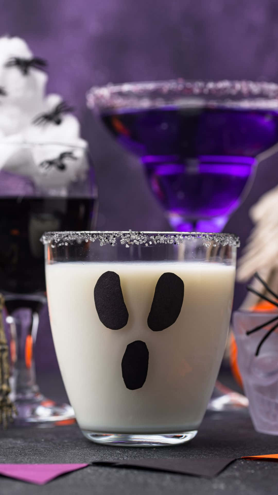 Spooky Cocktails 桌布 壁紙 — 一杯帶有糖邊的幽靈臉忌廉雞尾酒，周圍環繞著萬聖節主題飲品和裝飾。