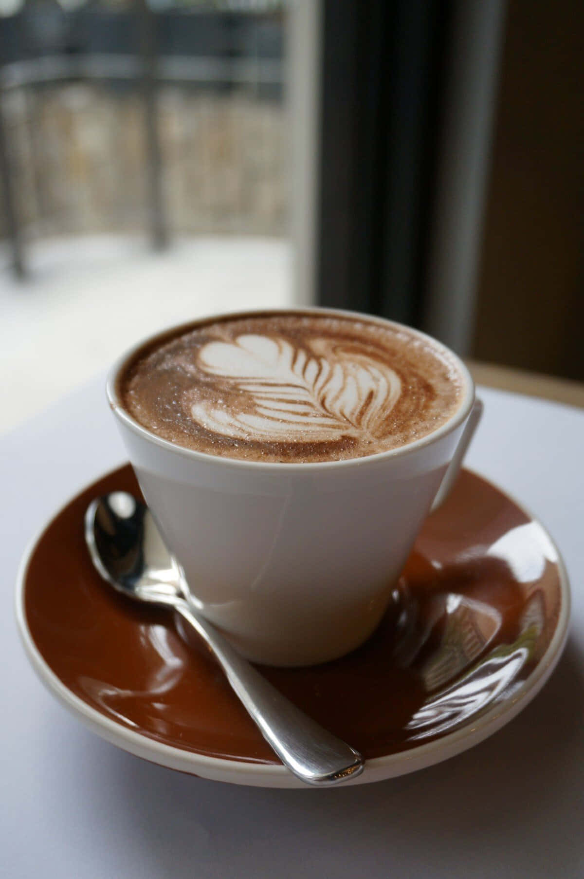 Coffee Cup 桌布 壁紙 — 一杯帶有葉子圖案嘅白色拉花咖啡，放喺棕色碟上，旁邊有隻匙羹，靠近窗邊。