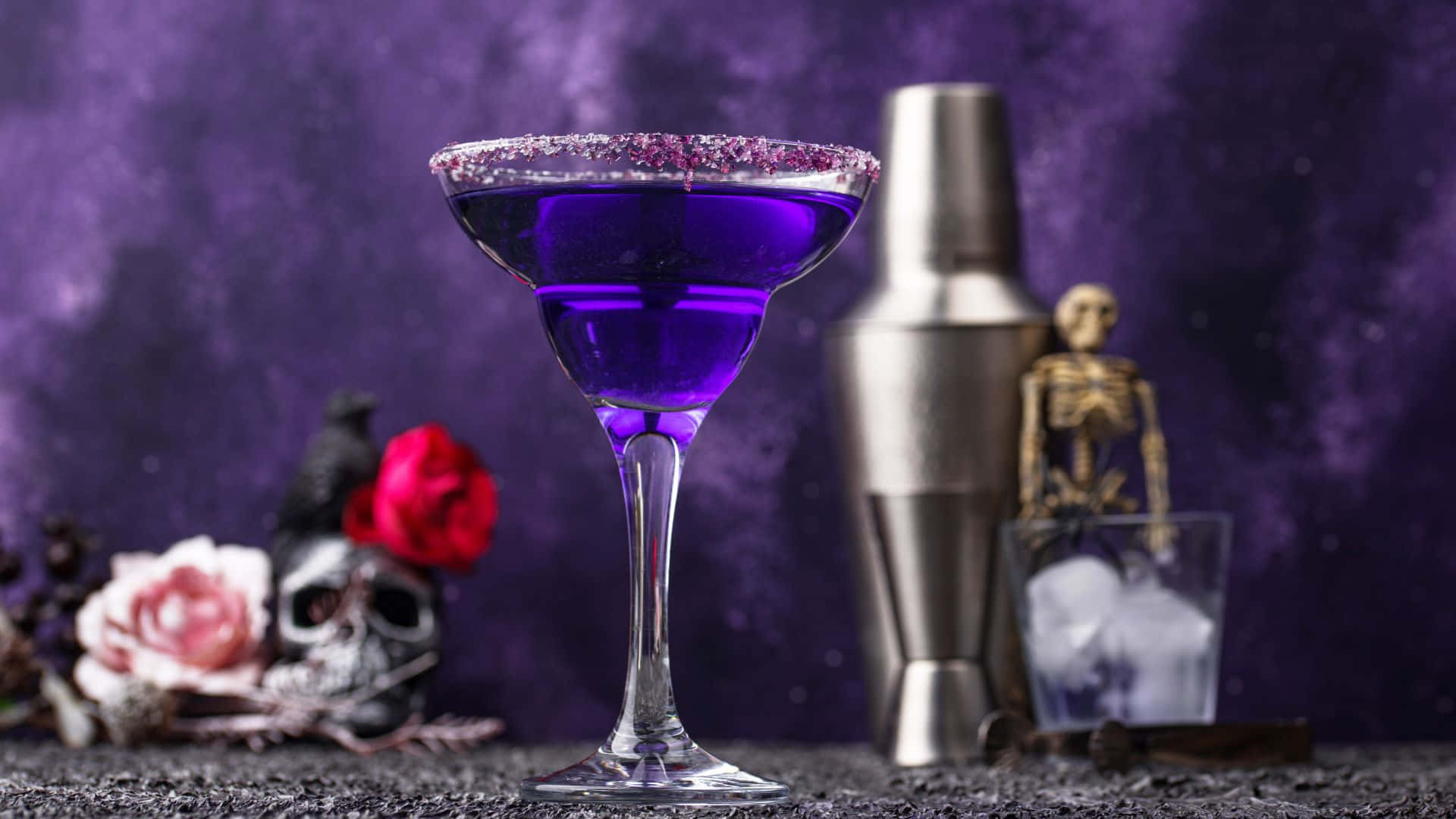 Spooky Cocktails 桌布 壁紙 — 一杯帶有閃粉邊的鮮豔紫色雞尾酒，旁邊放著雞尾酒調酒器和萬聖節主題裝飾，背景為深紫色。