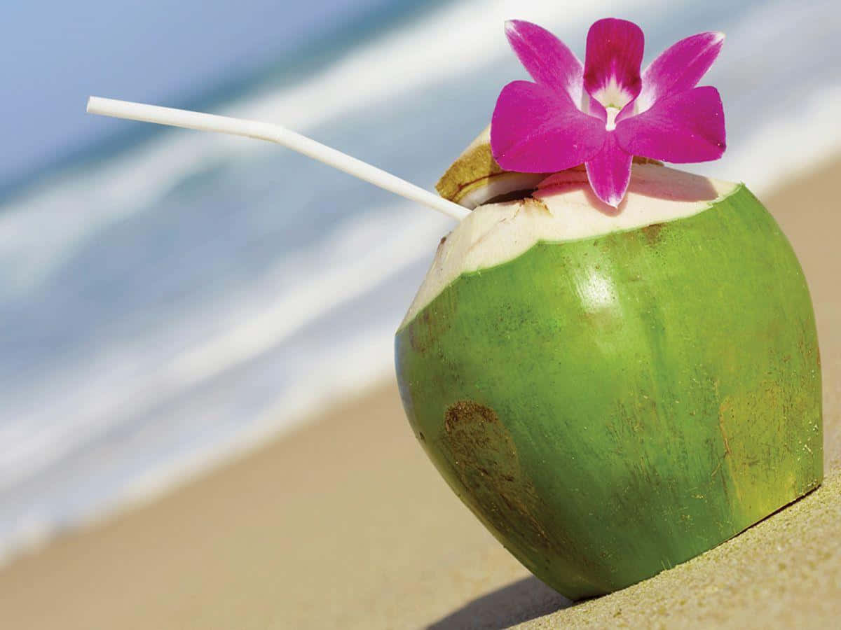 Coconut 桌布 壁紙 — 一杯新鮮的綠色椰子飲品，配上粉紅色蘭花和吸管，放在沙灘上，背景有海浪。