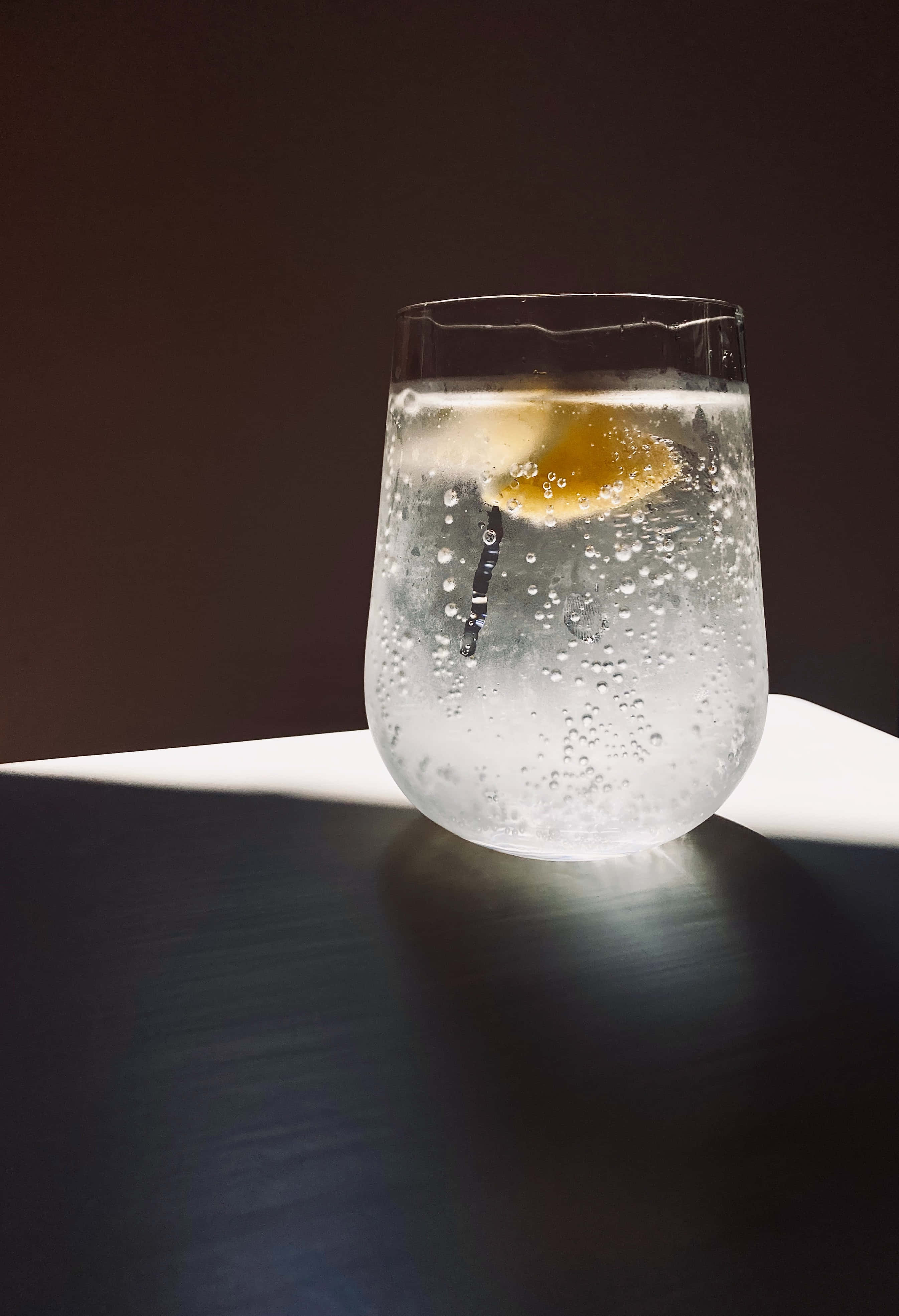 Hydration 桌布 壁紙 — 一杯梳打水，配上檸檬片和飲管，在深色桌子上沐浴在明亮的陽光中。