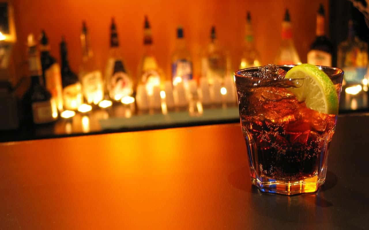alcohol 桌布 壁紙 — 一杯深琥珀色的雞尾酒，配上冰塊和青檸裝飾，在金色環境燈光下於吧檯上溫暖地閃耀。