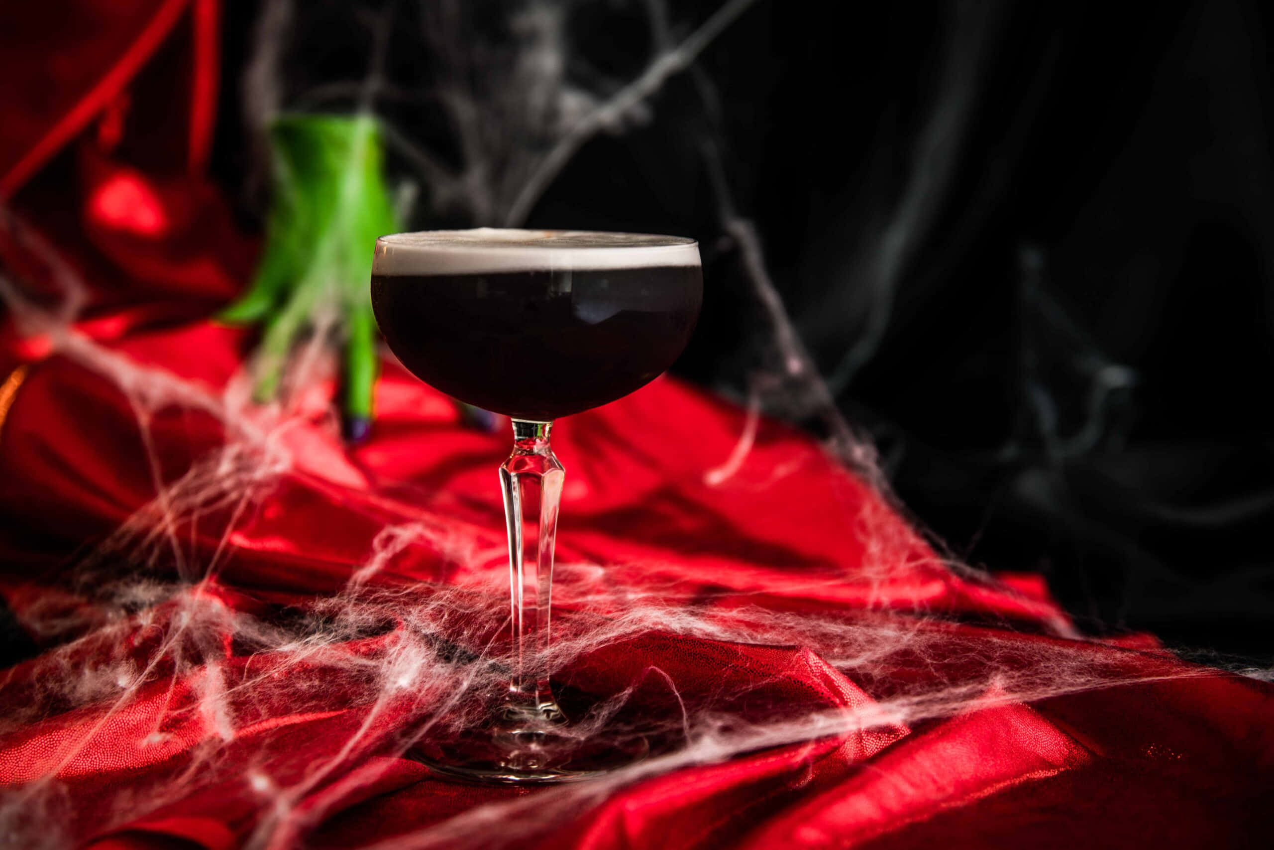 Halloween Cocktails 桌布 壁紙 — 一杯深色雞尾酒放在紅色布料上，布料上裝飾著白色蜘蛛網，背景中有一隻綠色女巫的手。