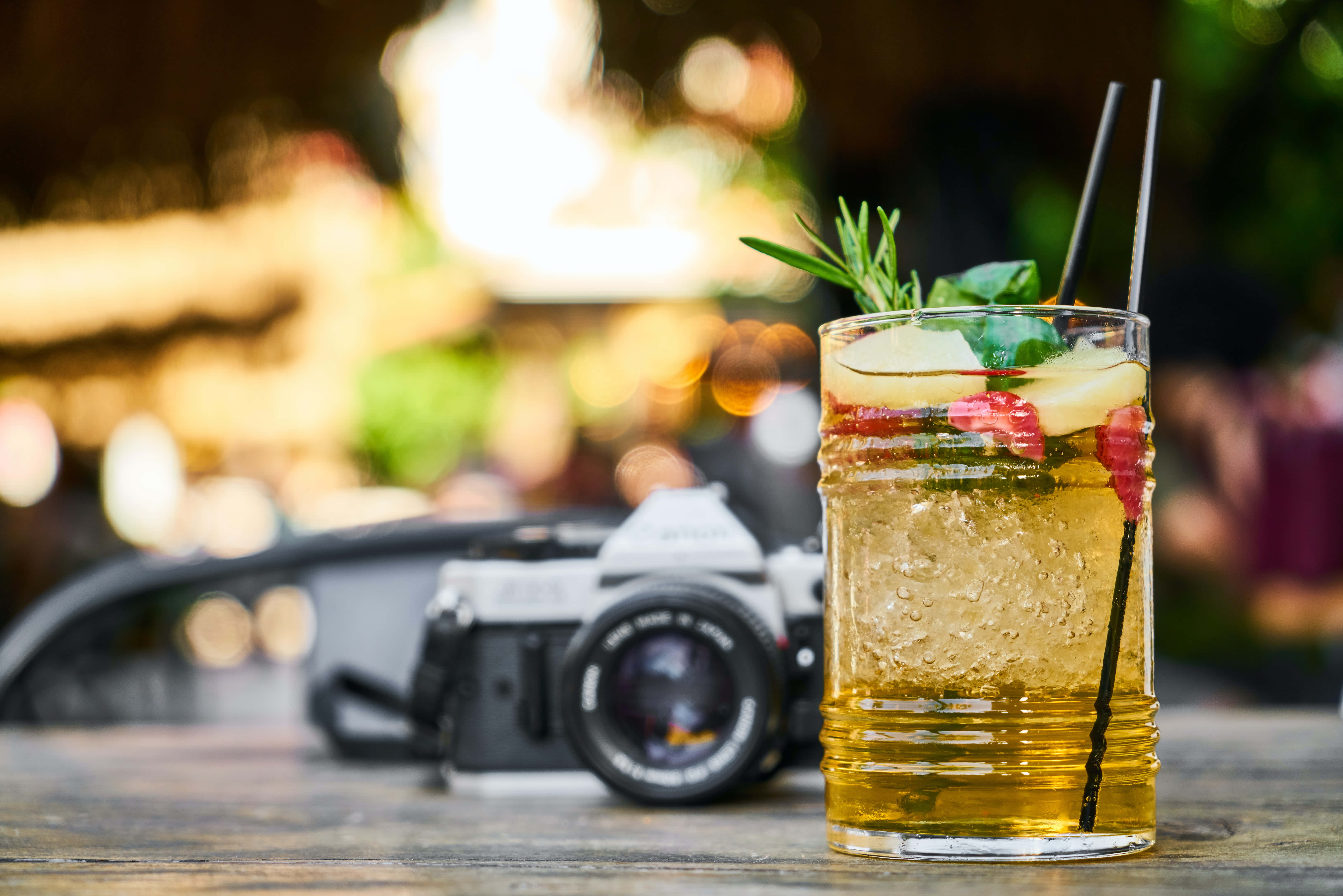 Summer Drinks 桌布 壁紙 — 一杯清爽嘅金色雞尾酒，用迷迭香同水果裝飾，放喺一張木枱上，旁邊有一部復古相機，背景係燈光溫暖嘅戶外酒吧。