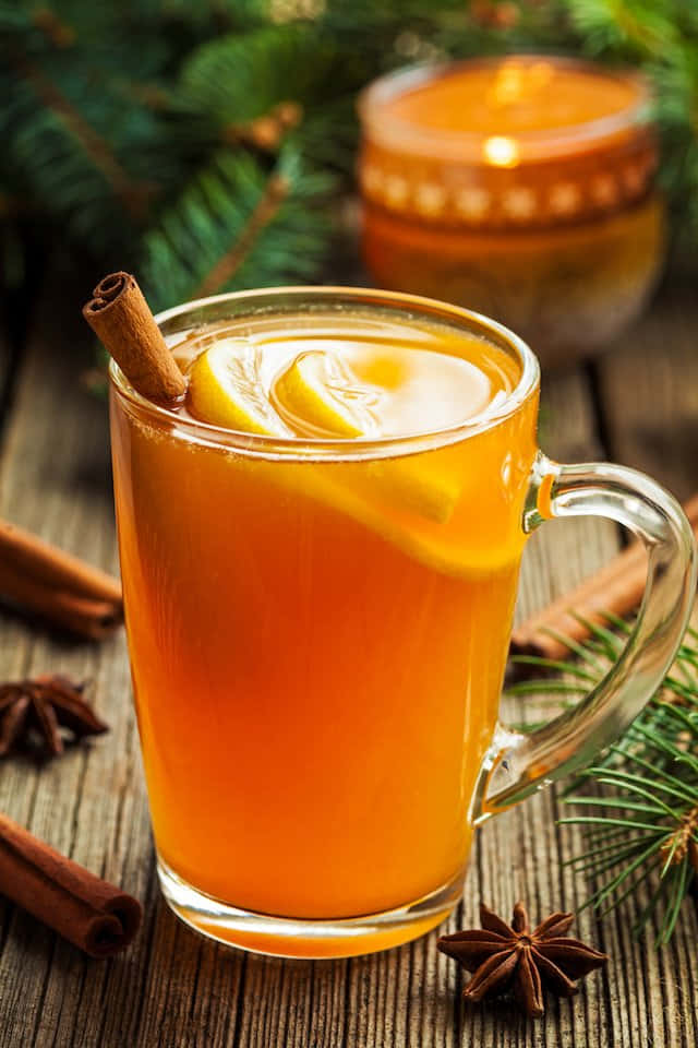 Winter Drinks 桌布 壁紙 — 一杯溫暖琥珀色香料飲品盛載於玻璃杯中，以肉桂棒和檸檬片裝飾，放在質樸木桌上，旁邊有松針和八角。