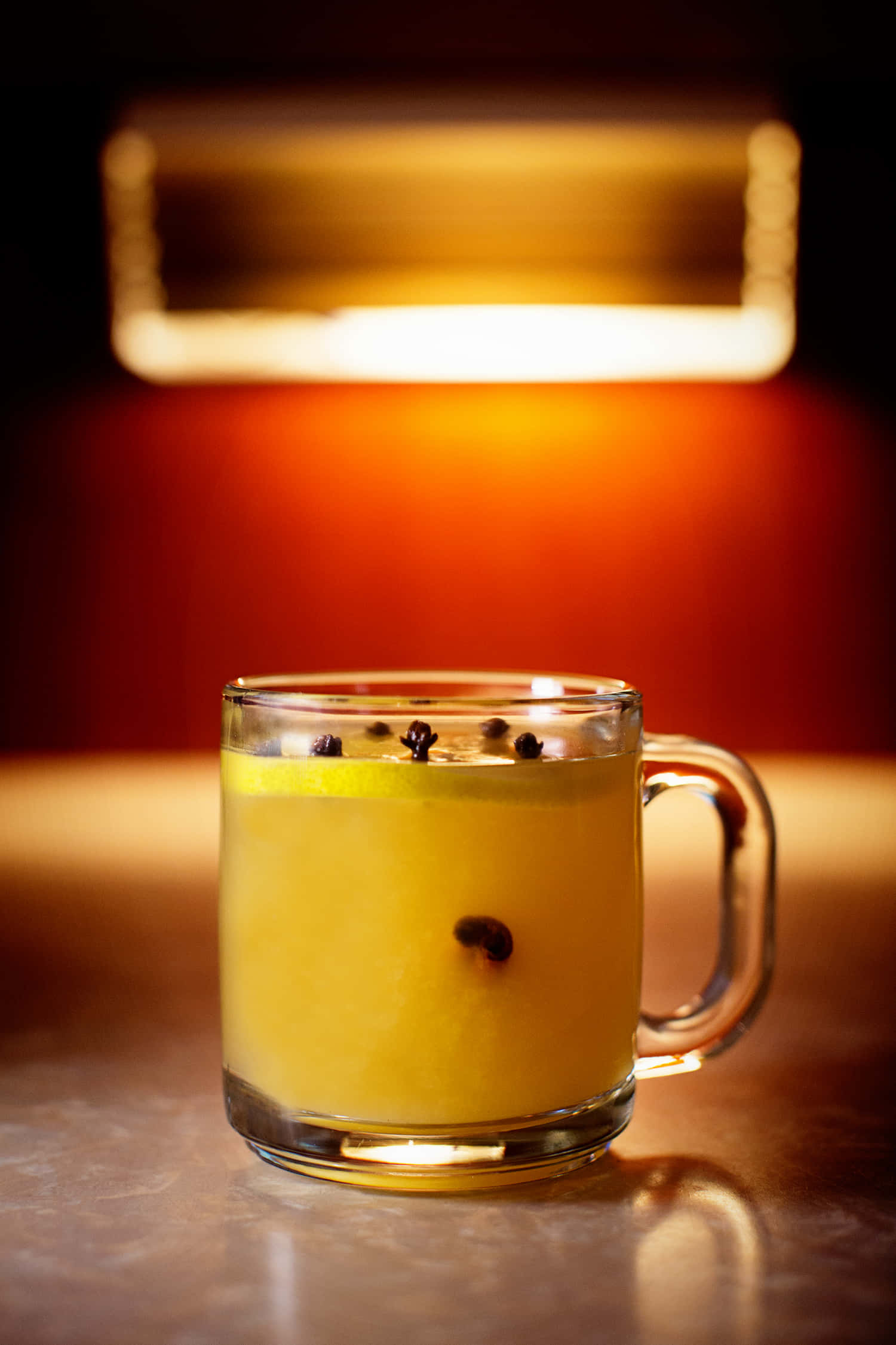 Winter Drinks 桌布 壁紙 — 一杯溫暖的琥珀色飲品，漂浮著香料，在舒適的室內環境中，在柔和的天花燈光下閃耀。