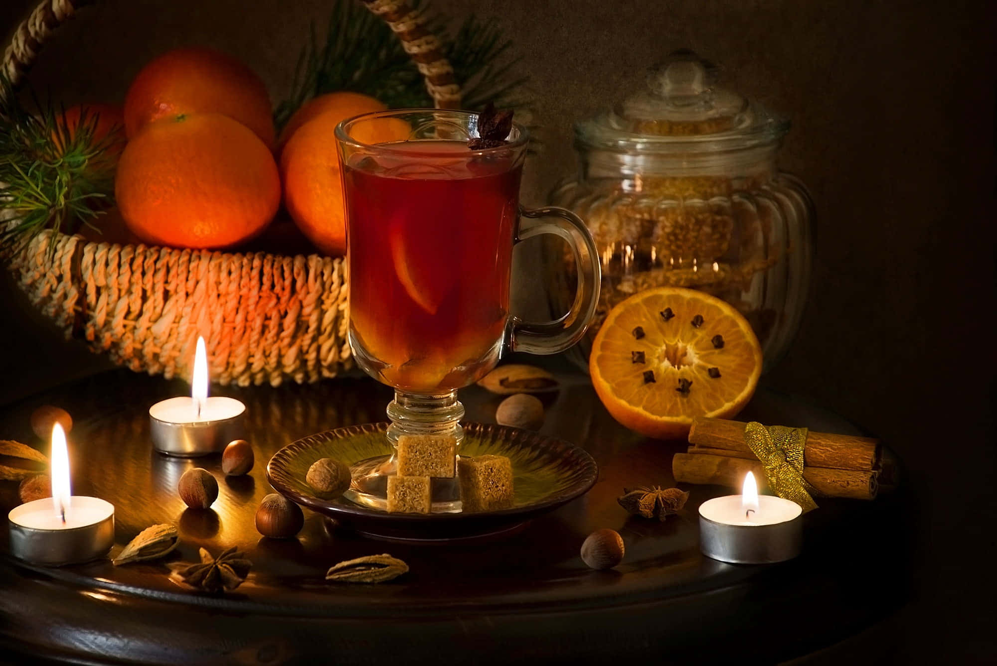 Mulled Wine 桌布 壁紙 — 一杯溫暖的香料茶在橙子、肉桂和點燃的蠟燭旁閃耀，構成舒適的冬季靜物畫。