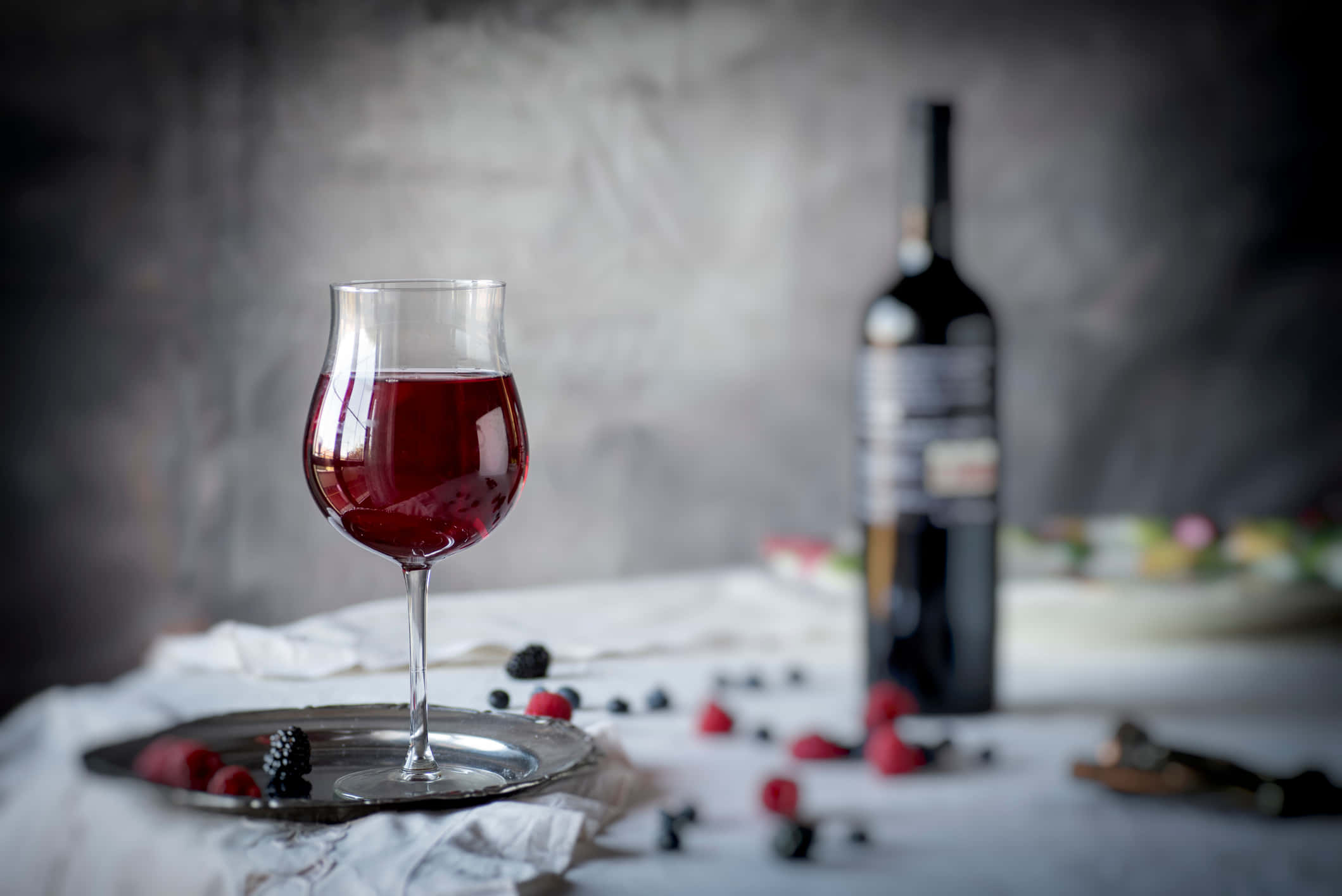 Blackberry Wine 桌布 壁紙 — 一杯濃郁的紅酒放在銀托盤上，旁邊有一瓶酒和散落的漿果，置於有紋理的白色布料上，背景為柔和的灰色。