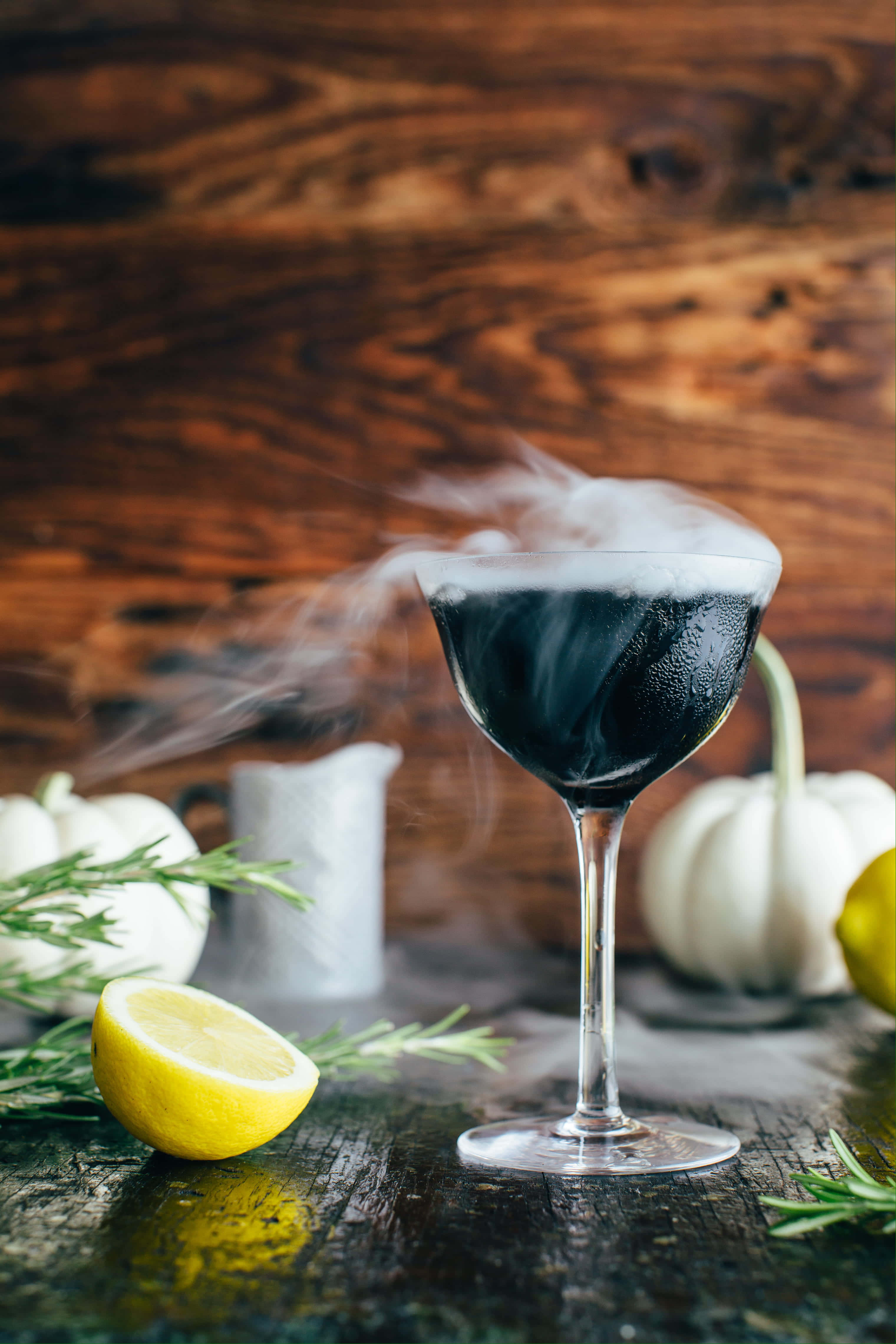 Halloween Drinks 桌布 壁紙 — 一杯煙燻黑色雞尾酒放在高腳杯裡，置於鄉村風木桌上，周圍有白色南瓜、檸檬和迷迭香。