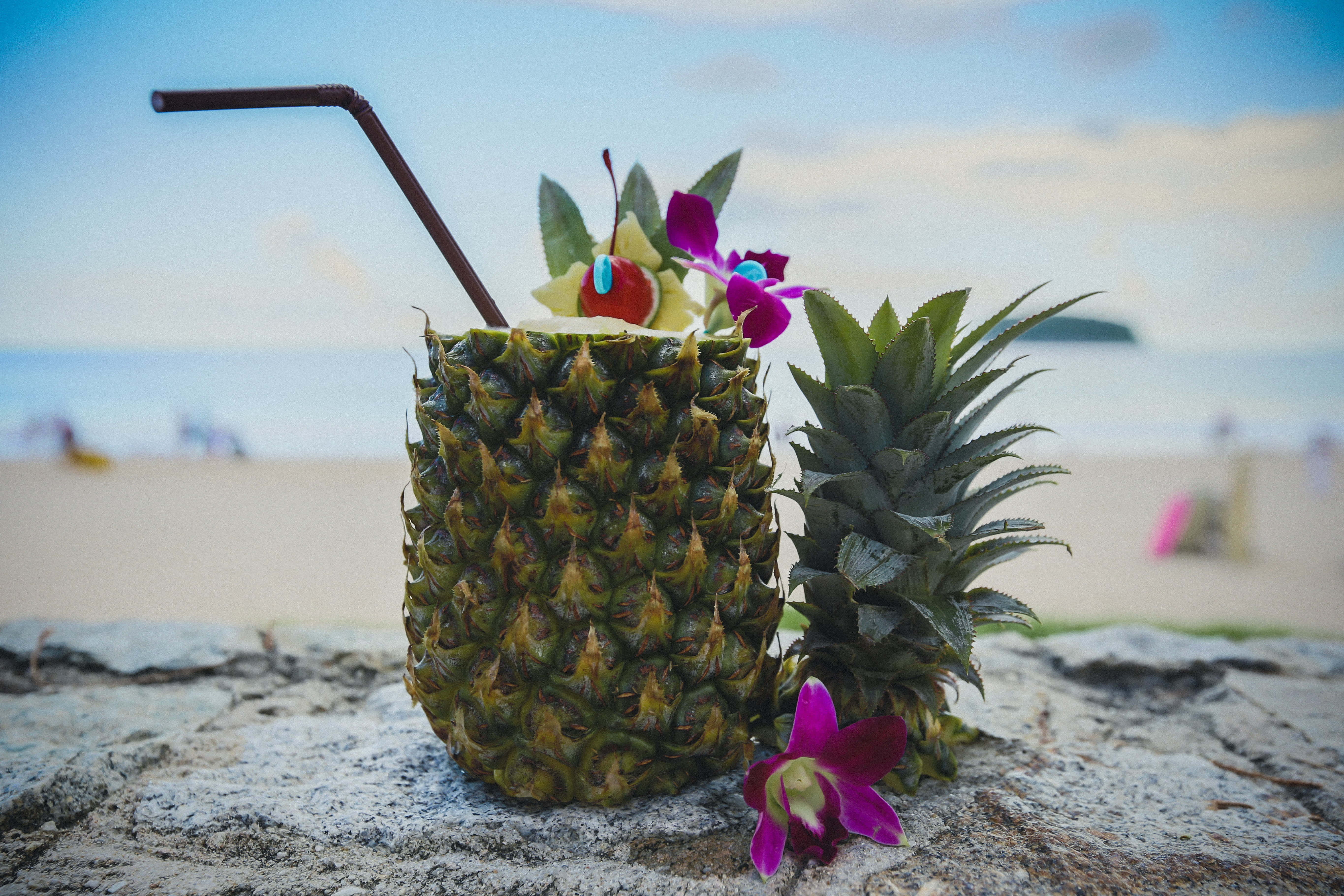 Summer Drinks 桌布 壁紙 — 一杯熱帶菠蘿飲品，飾以花朵和櫻桃，放在陽光海灘的石頭上，背景是海洋。