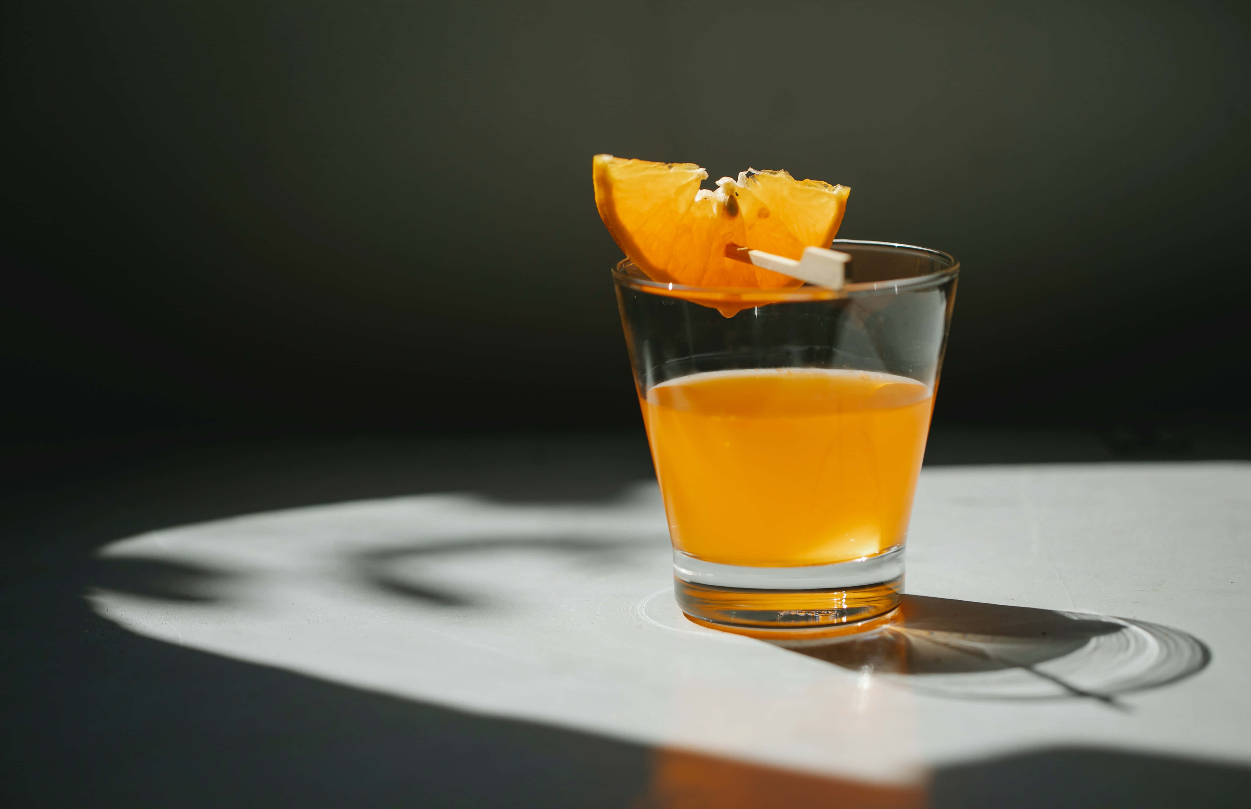 Summer Drinks 桌布 壁紙 — 一杯琥珀色雞尾酒，飾以橙片，置於白色表面上，在聚光燈下，背景為深色。