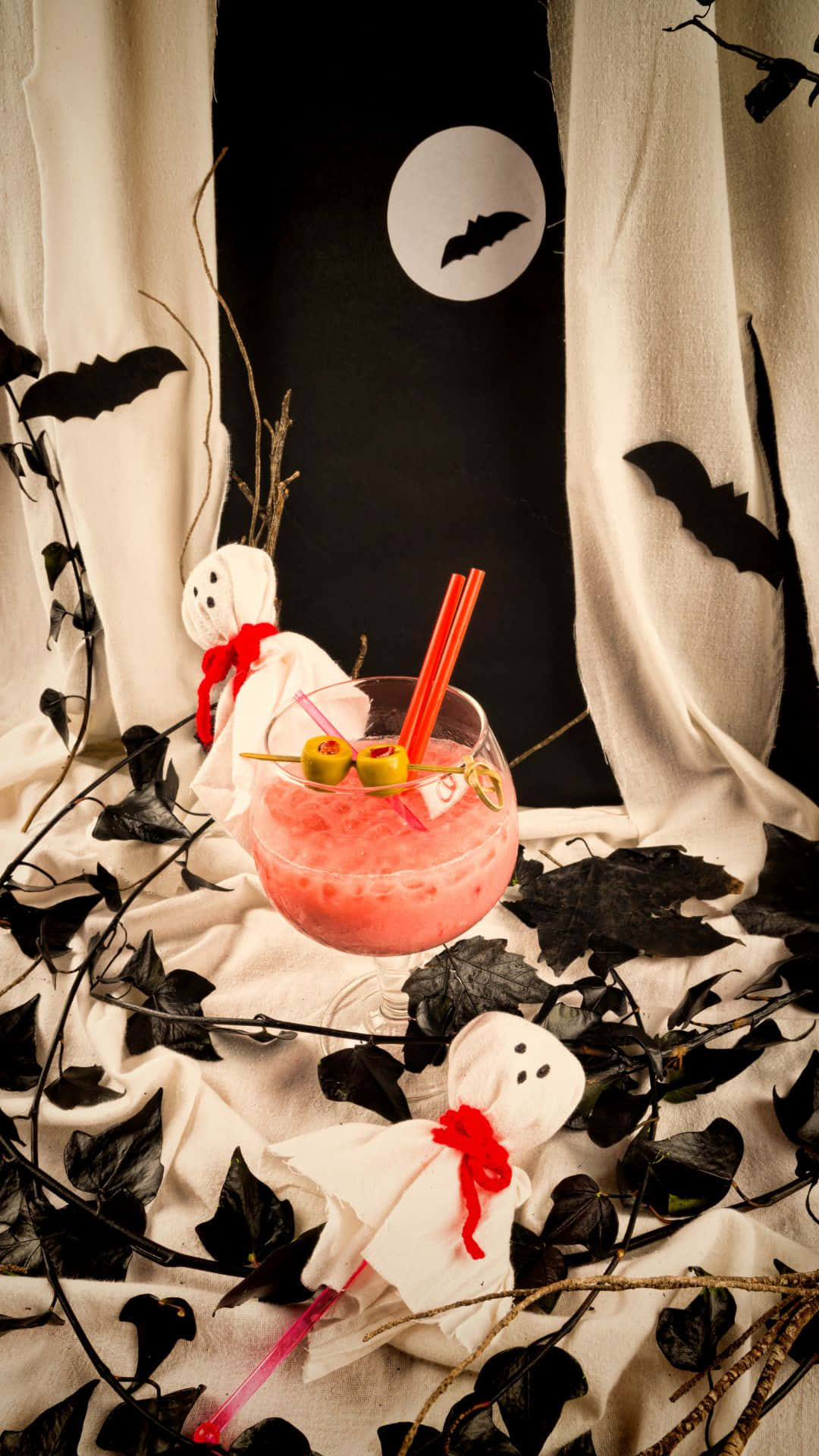 Spooky Cocktails 桌布 壁紙 — 一杯粉紅色萬聖節雞尾酒（配橄欖和飲管）放在蝙蝠月亮背景下的鬼怪裝飾旁。