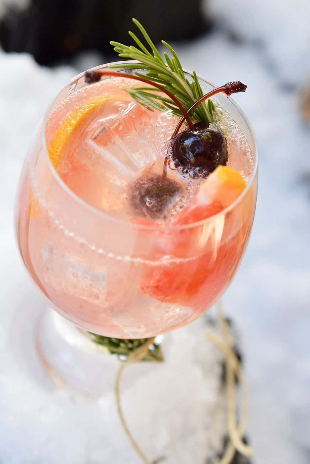 Winter Drinks 桌布 壁紙 — 一杯粉紅色雞尾酒，配有冰塊、橙片、櫻桃和迷迭香裝飾，放在雪地上。