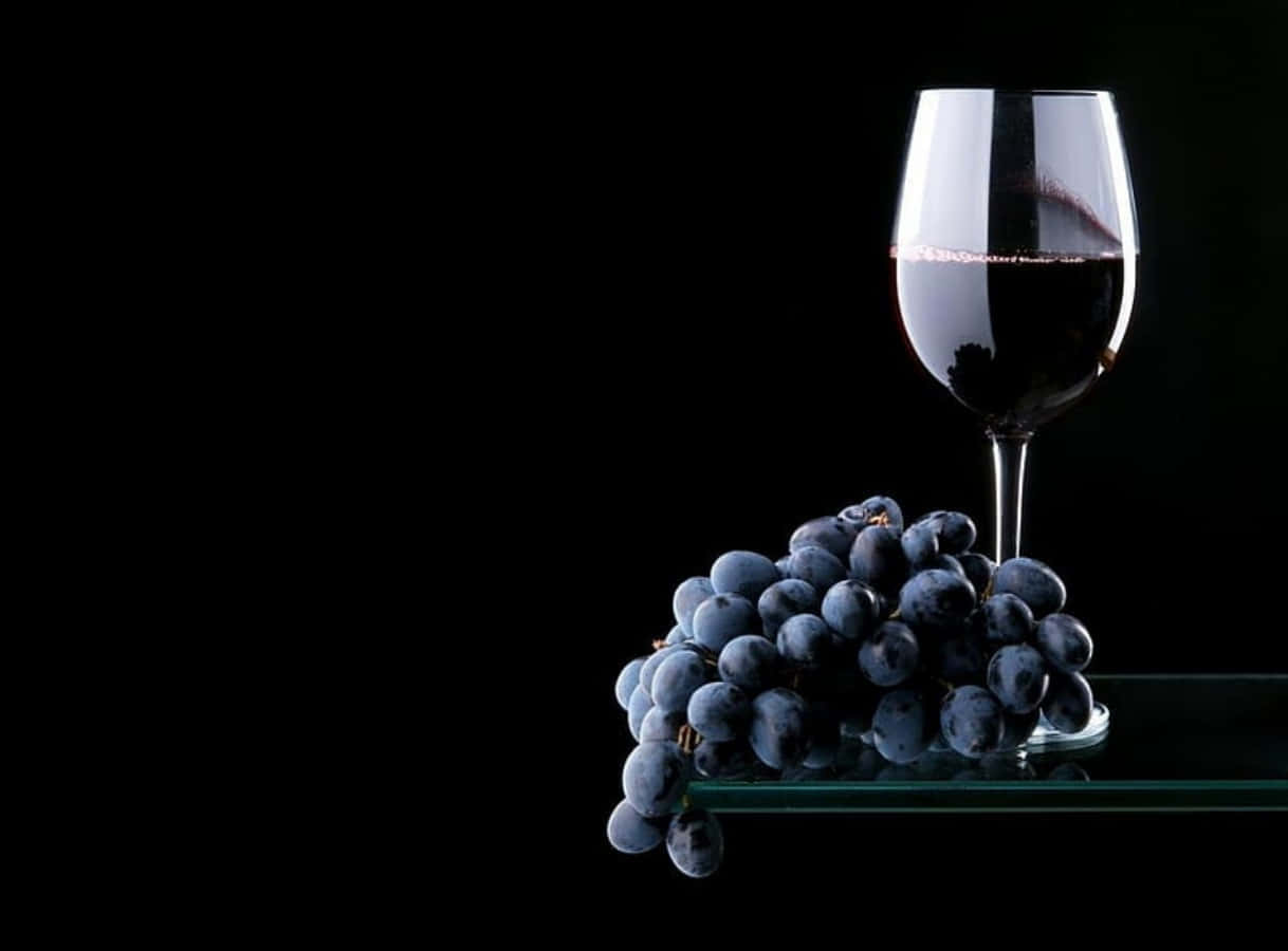 Black Wine 桌布 壁紙 — 一杯紅酒和一串深色葡萄優雅地放在玻璃表面上，背景是純黑色。