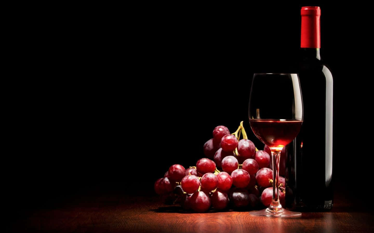 Red Wine Glass 桌布 壁紙 — 一杯紅酒和一瓶紅酒放在深色木桌上，旁邊是一串成熟的紅葡萄，背景為黑色。