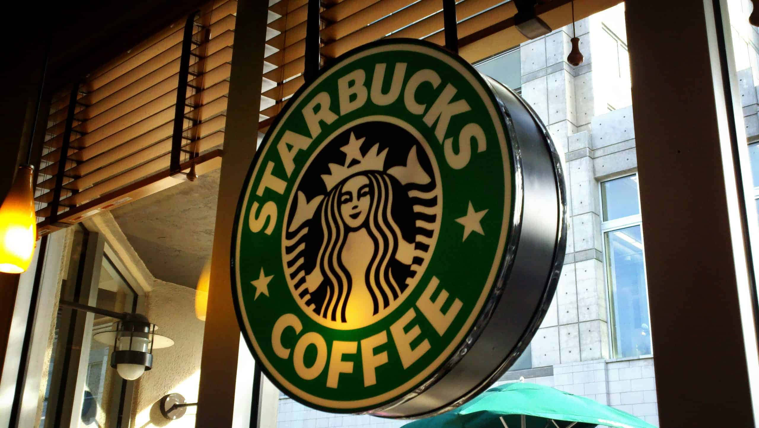 一杯經典的星巴克咖啡，為完美早晨開幕 壁紙 — 一個發光的Starbucks咖啡標誌掛在店舖外，店內有溫暖的燈光和木製百葉窗。