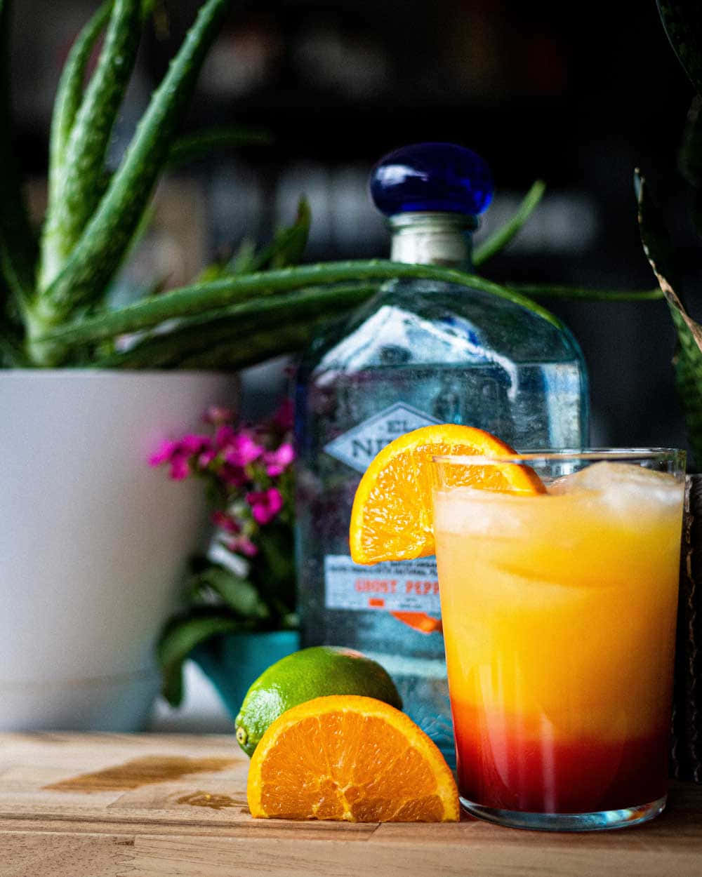 Beautiful Drink 桌布 壁紙 — 一杯色彩鮮豔的漸變雞尾酒，配上冰塊和橙片，旁邊放著一瓶氈酒和新鮮柑橘，置於木桌上。