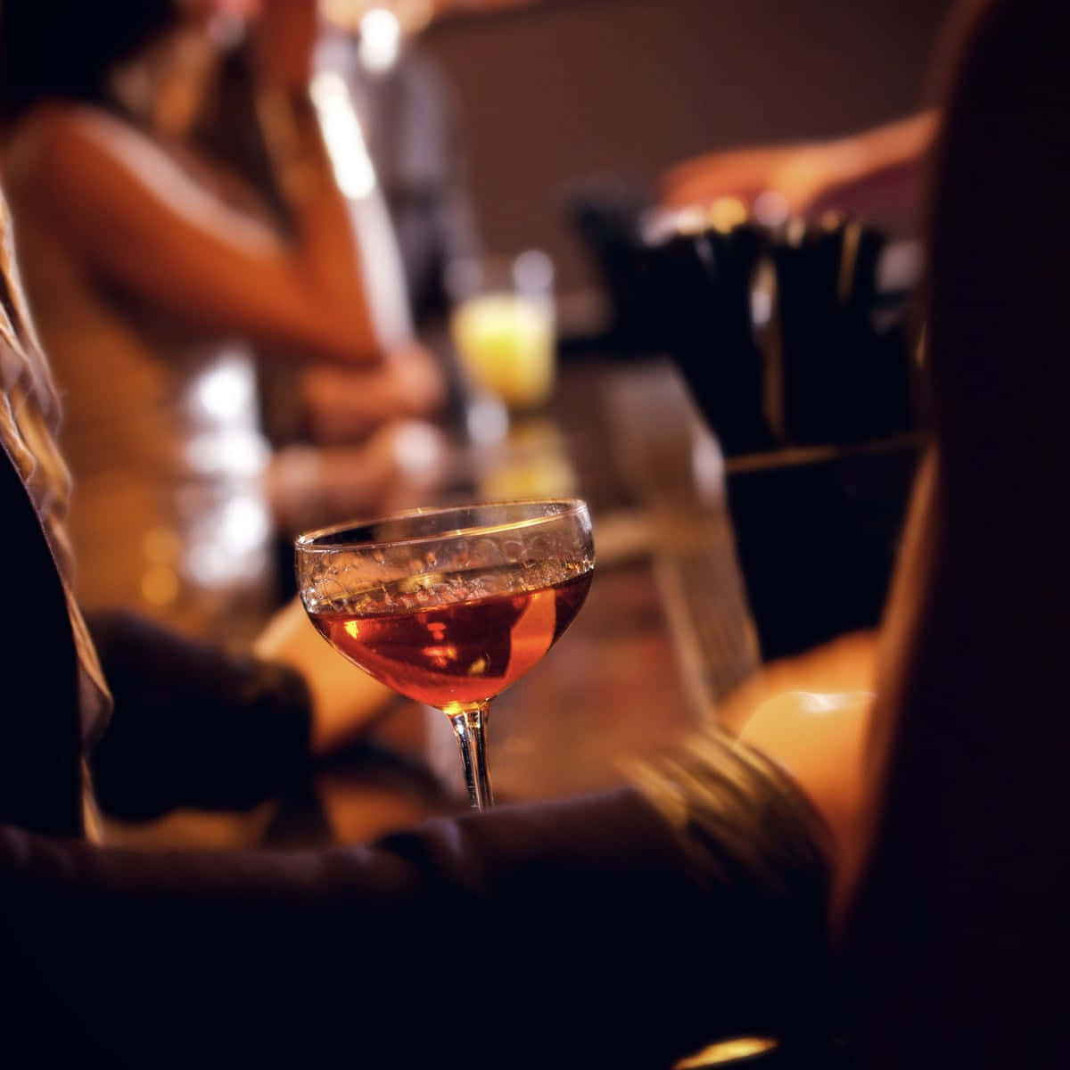 Drunk 桌布 壁紙 — 一杯裝滿琥珀色液體的雞尾酒杯特寫，背景是燈光昏暗的酒吧和模糊的顧客。
